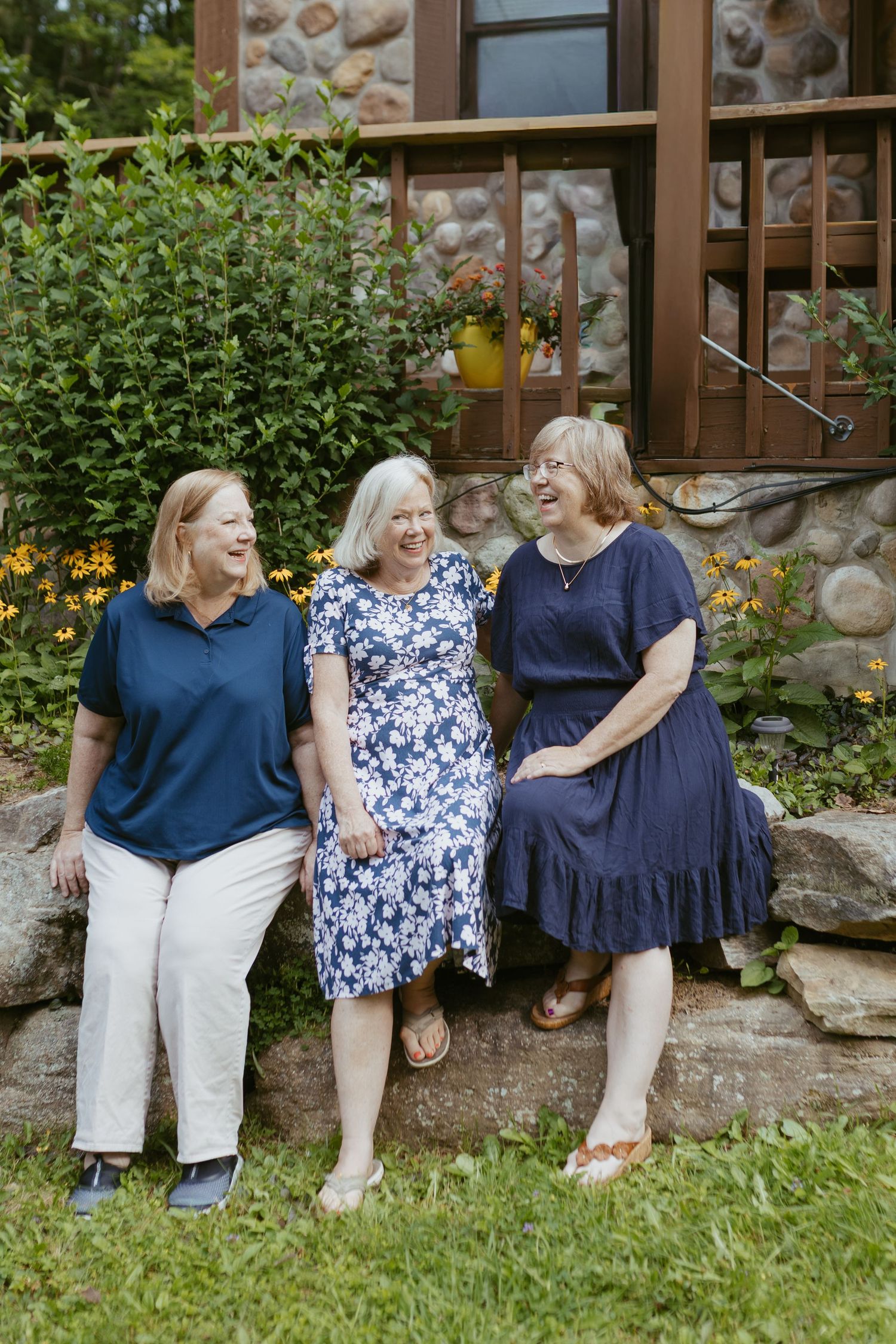 Three older women in blue outfits laughing together while sitting on a stone wall in a garden.