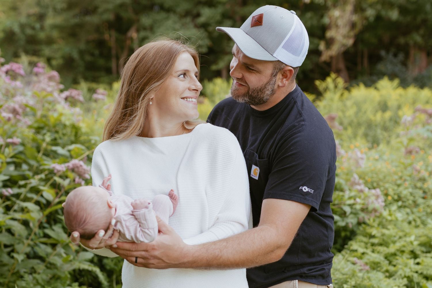 A couple smiles at each other while holding their newborn baby outdoors among lush green foliage and pink flowers.