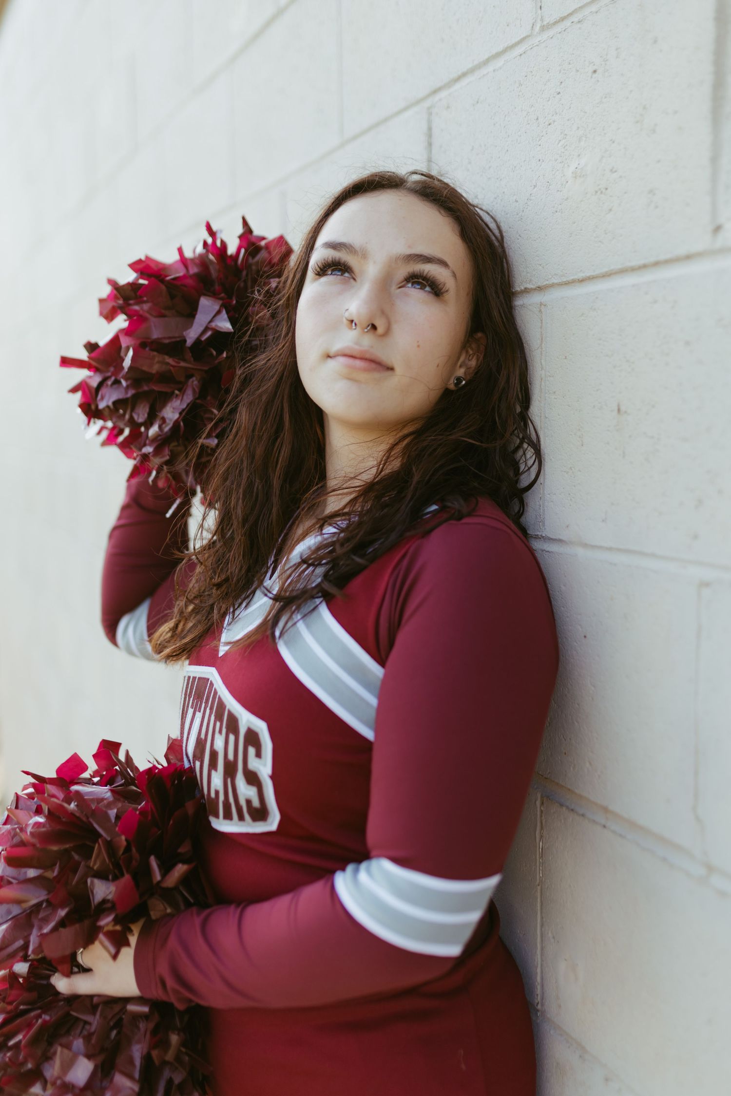 A young cheerleader in a maroon uniform holds pom-poms while leaning against a white brick wall, gazing upward.