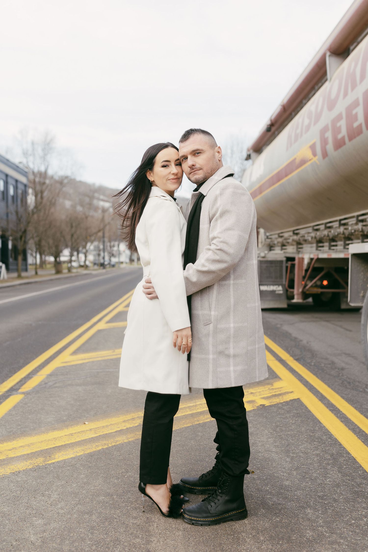 Stylish couple embracing on a city street, wearing elegant white and plaid coats with black pants outdoors.