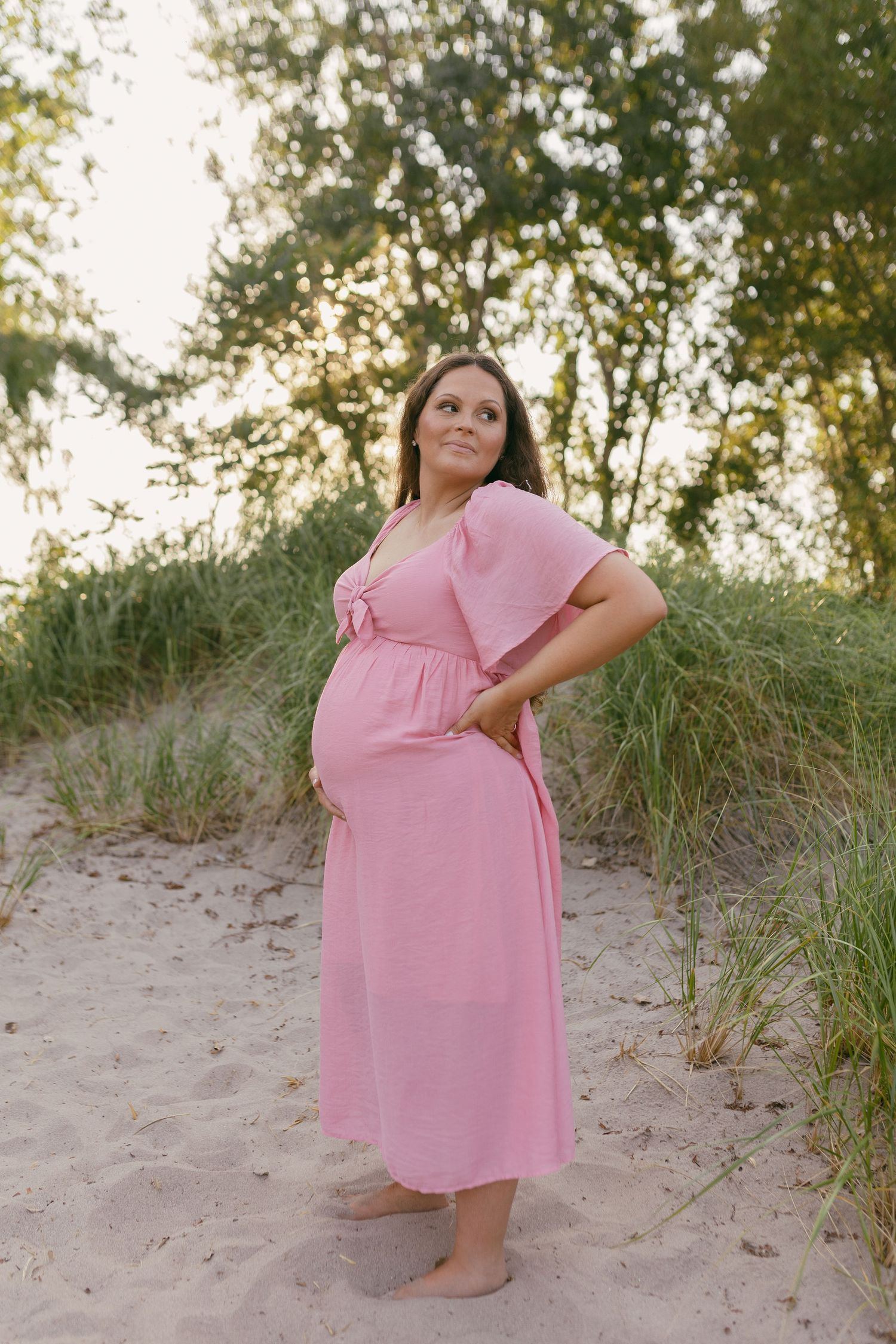 Maternity photo of woman in pink dress posing on sandy beach at sunset.