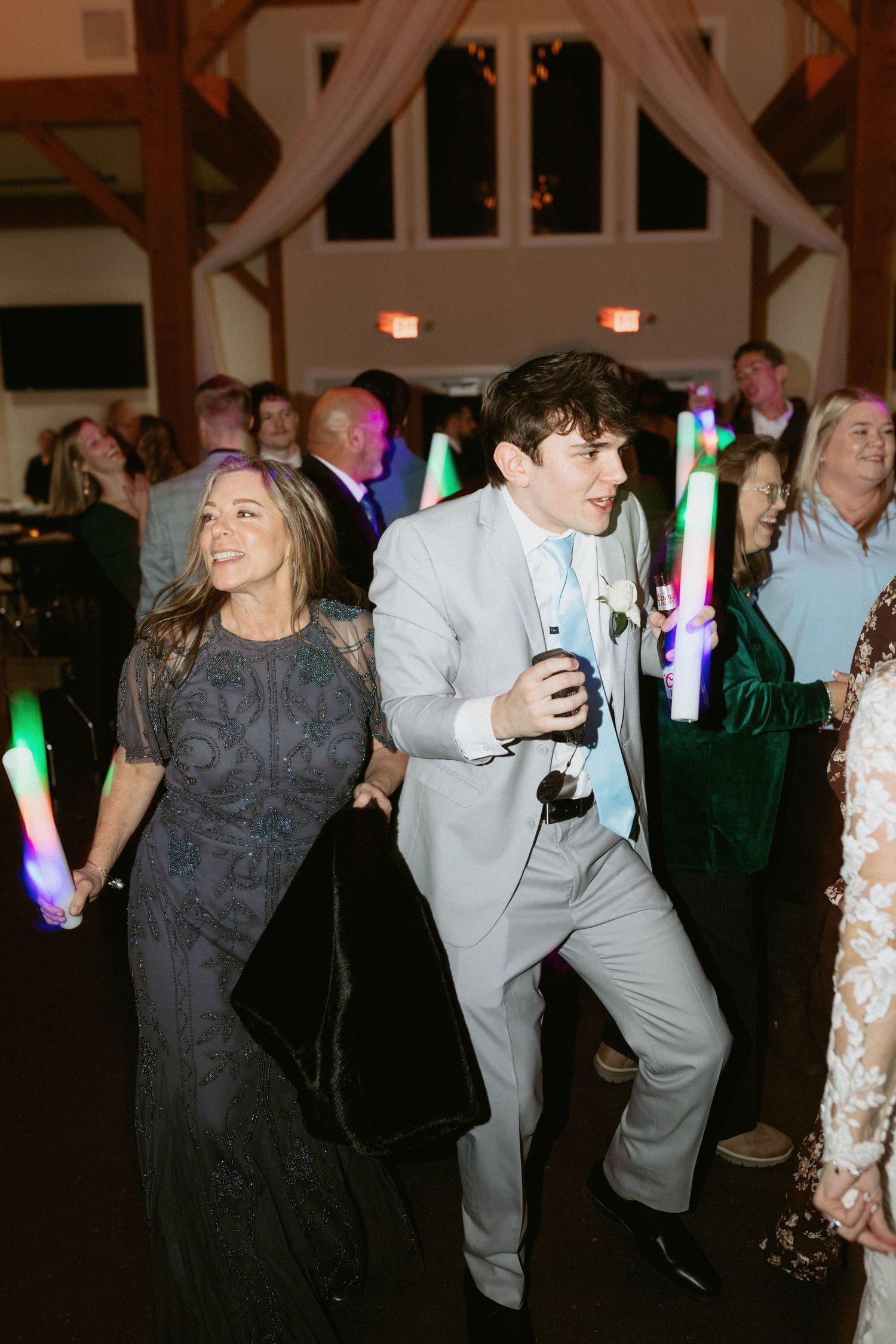 Wedding party dances energetically under colorful lights in a rustic barn-style venue.
