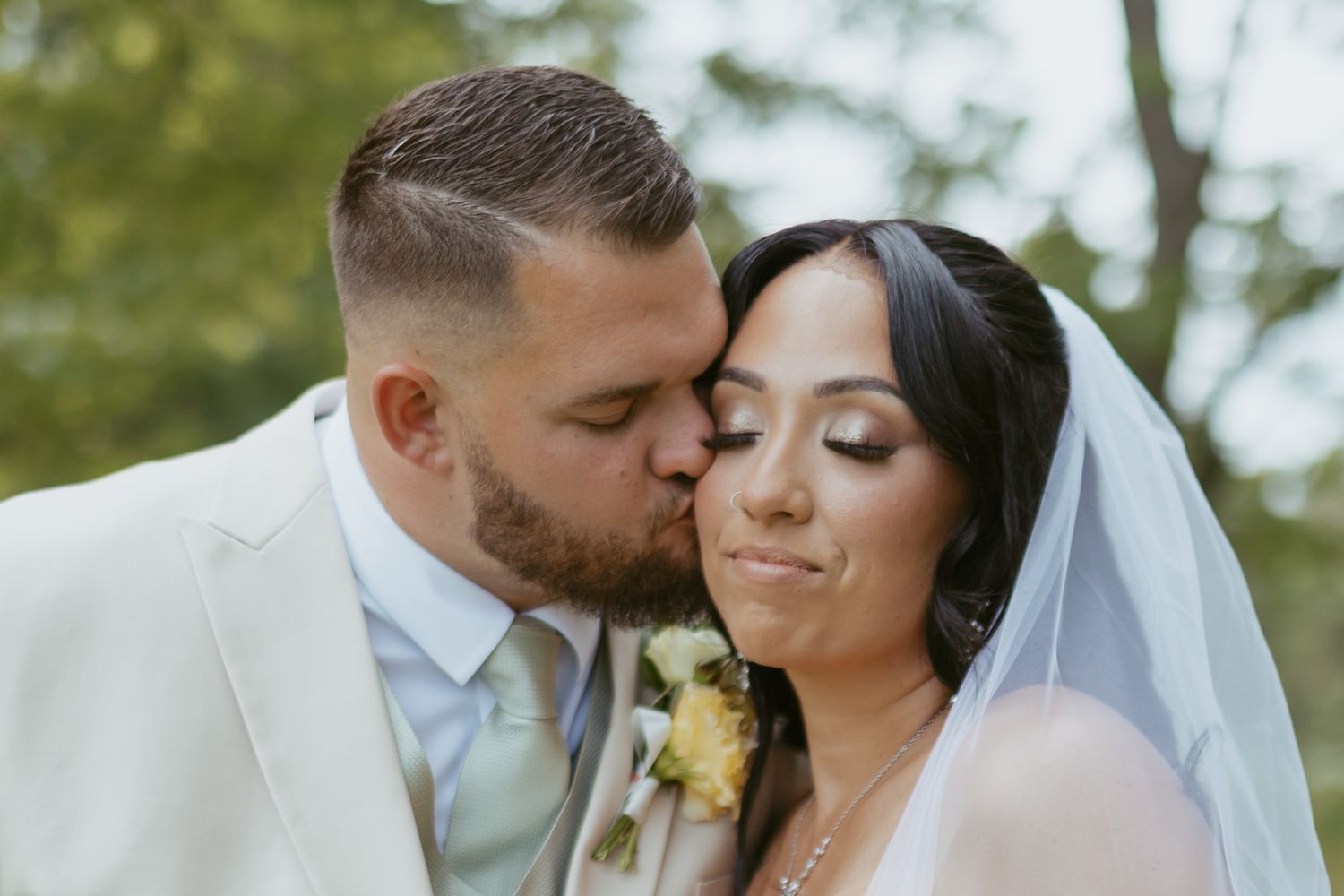 Groom in white suit kissing bride on cheek, bride wearing veil with yellow rose bouquet, outdoor wedding portrait.