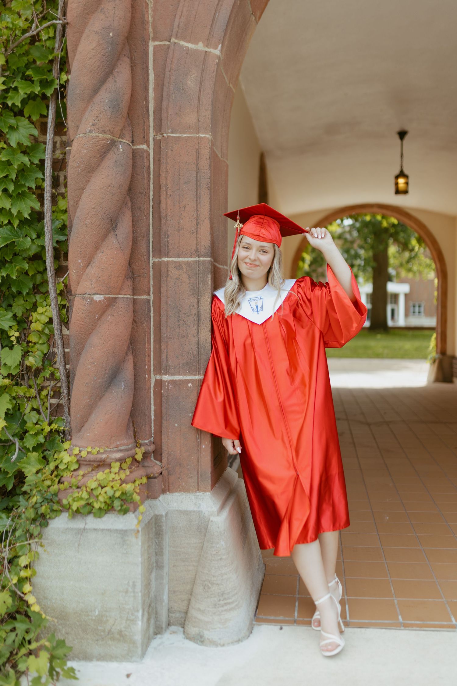 Young woman in red graduation cap and gown posing proudly by stone archway column on campus.