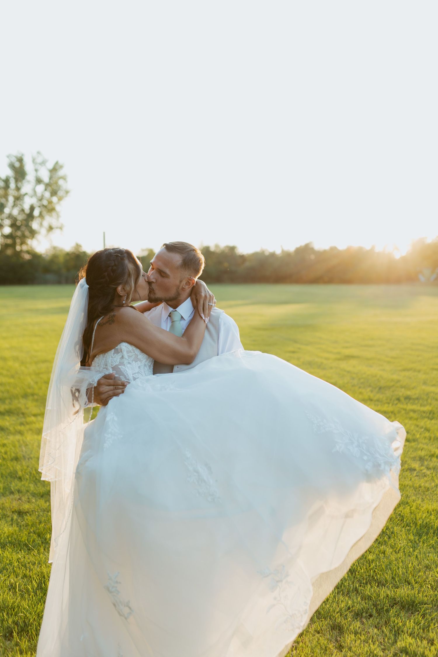 Groom carries bride in white gown and veil across green field at golden hour sunset.