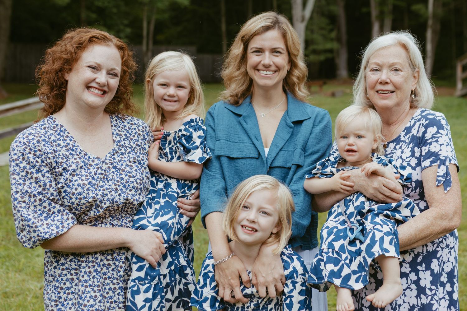 Multi-generational family of women and girls in matching blue floral dresses posing outdoors for a portrait.