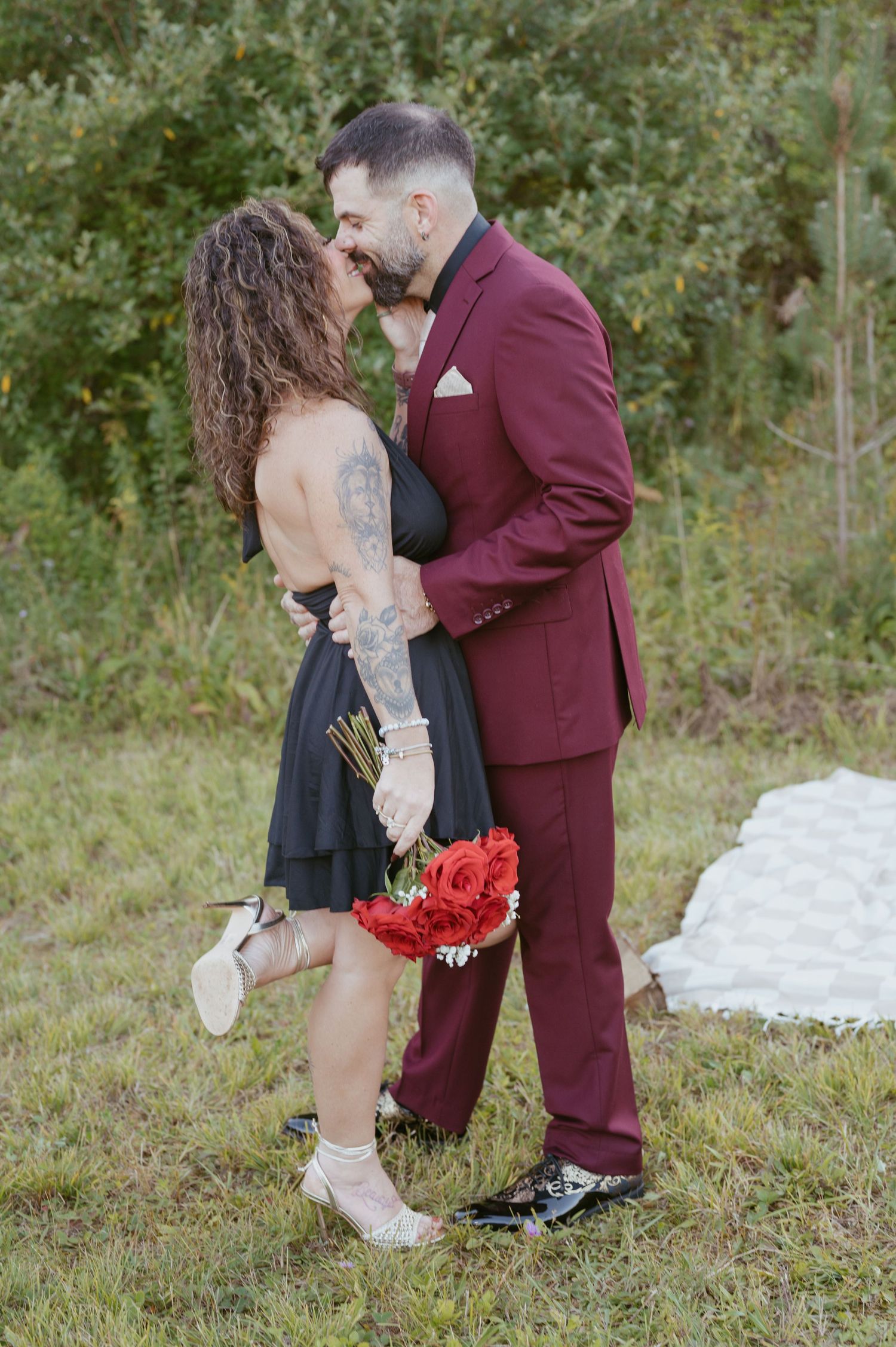 Couple kissing outdoors, woman in black dress holding red roses, man in burgundy suit, green trees background.
