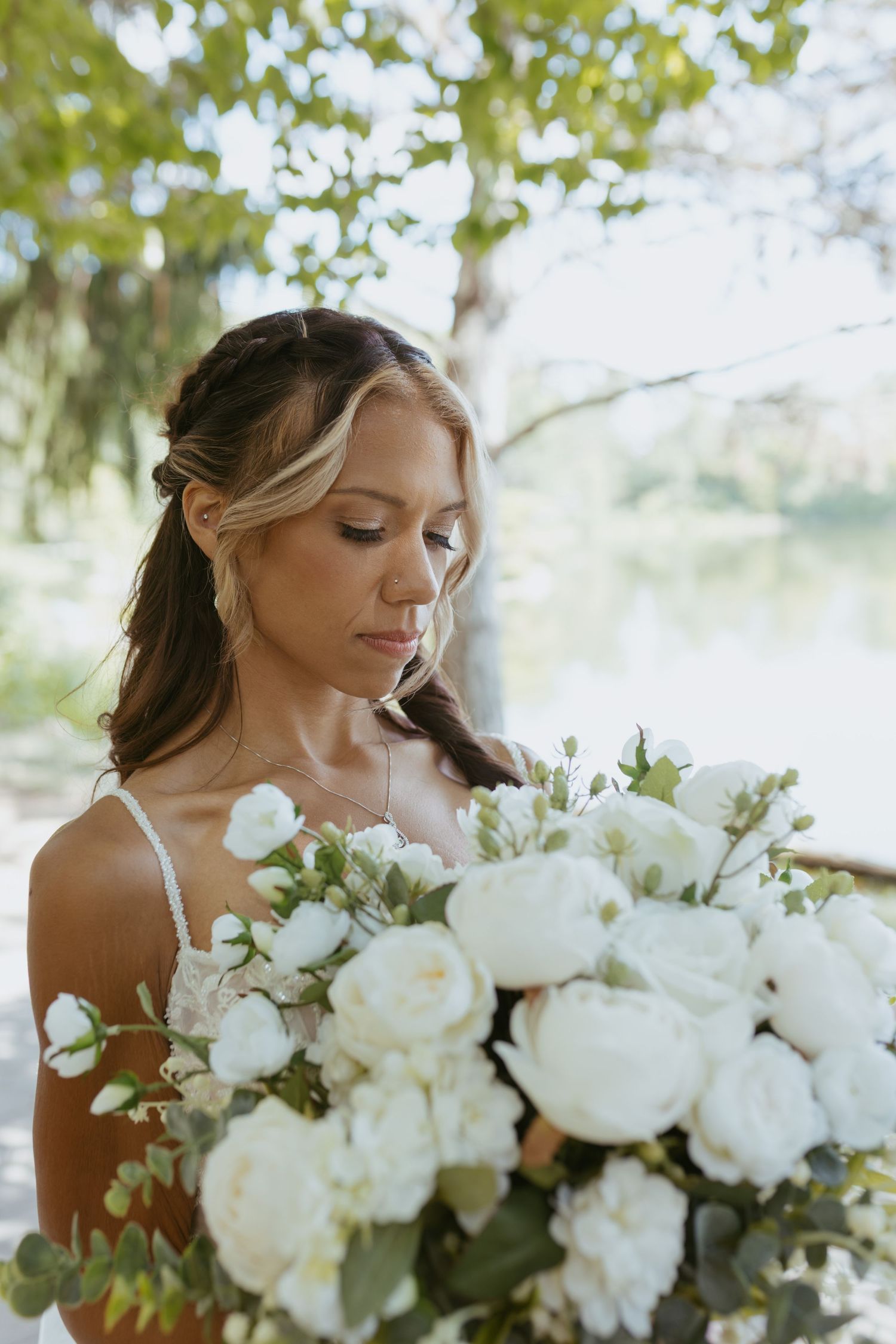 Bride in white dress gazes at large white peony bouquet outdoors near a serene lake surrounded by lush green trees.