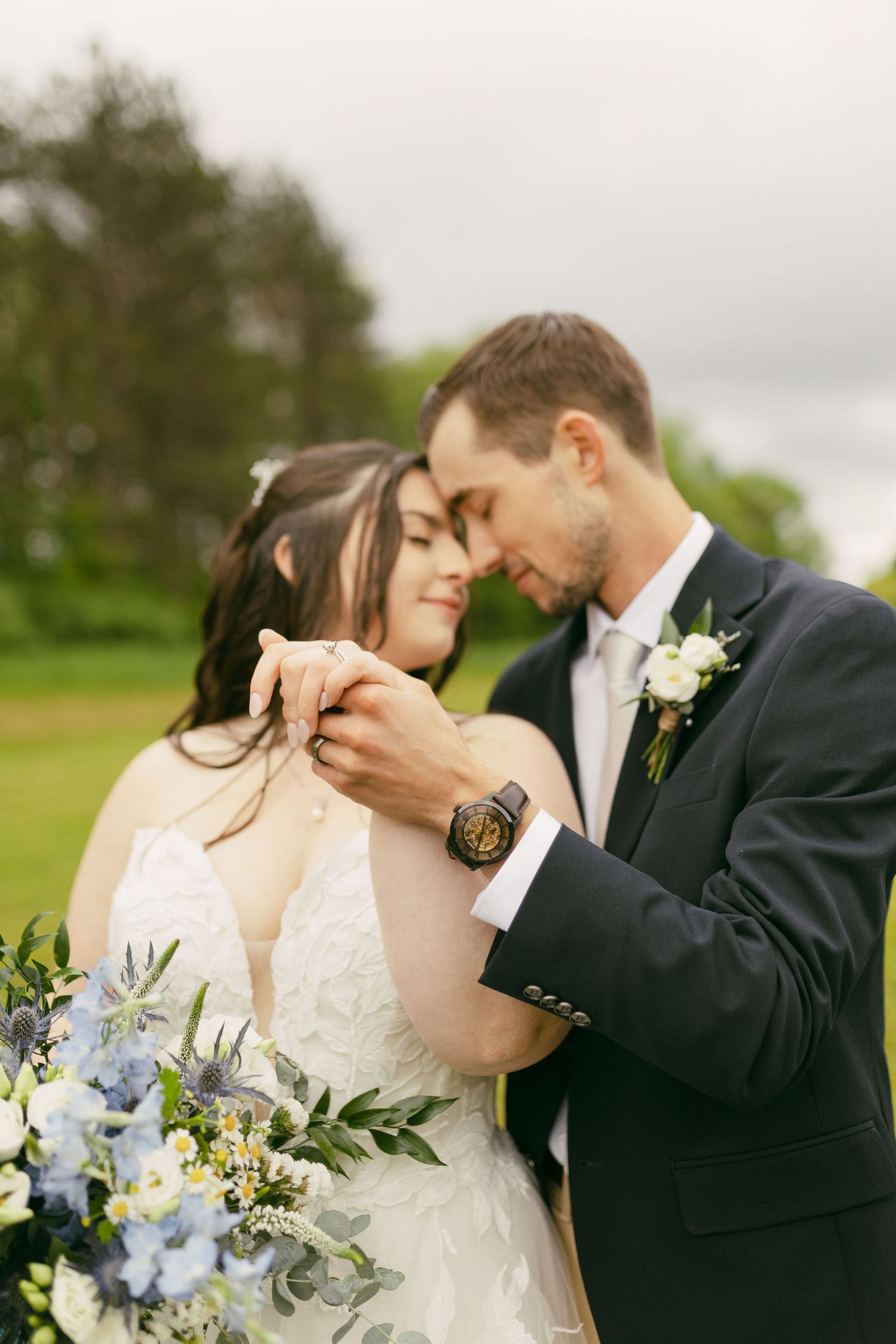Romantic wedding couple embraces in an outdoor setting with blue and white floral bouquet details.