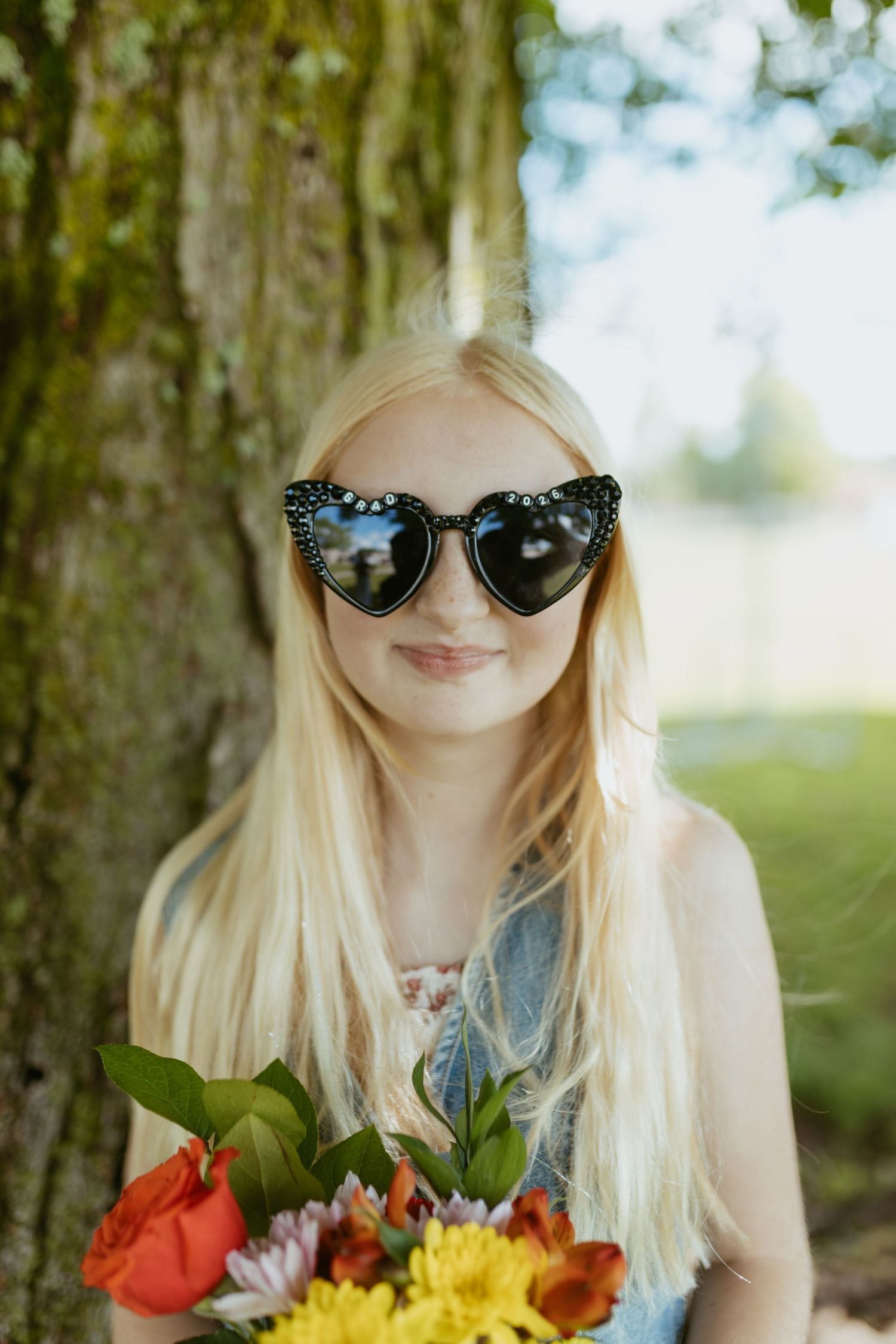 Blonde girl wearing heart-shaped cat-eye sunglasses holding colorful flowers outdoors near a tree.