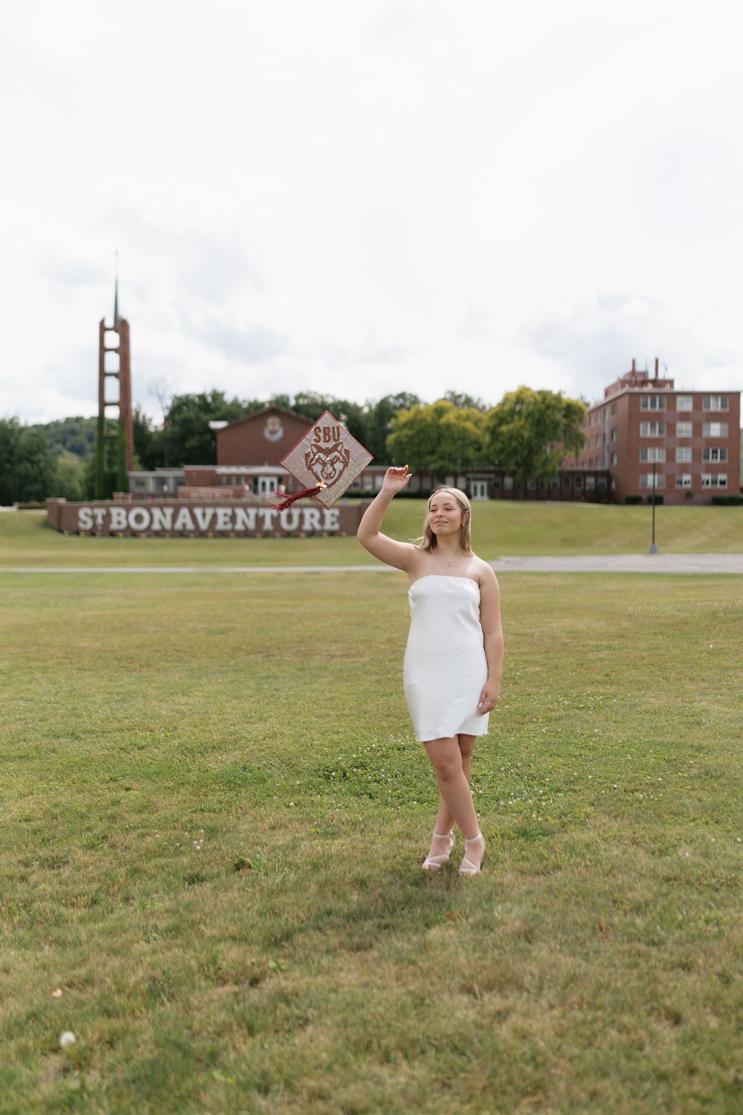 A young woman in a white strapless dress poses on green grass in front of the St. Bonaventure University sign.