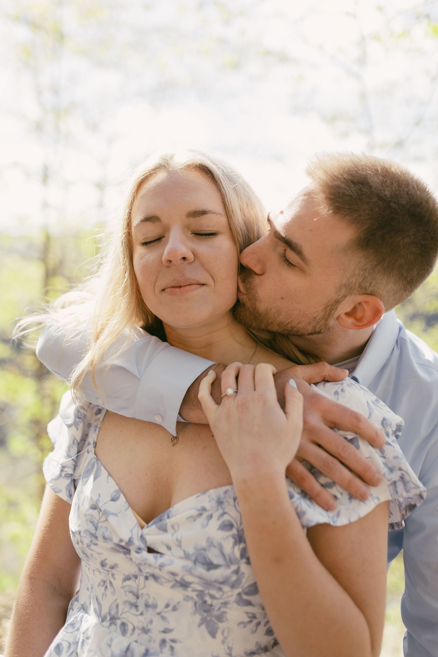 Man tenderly kissing woman on cheek outdoors in sunlit blossoming garden.