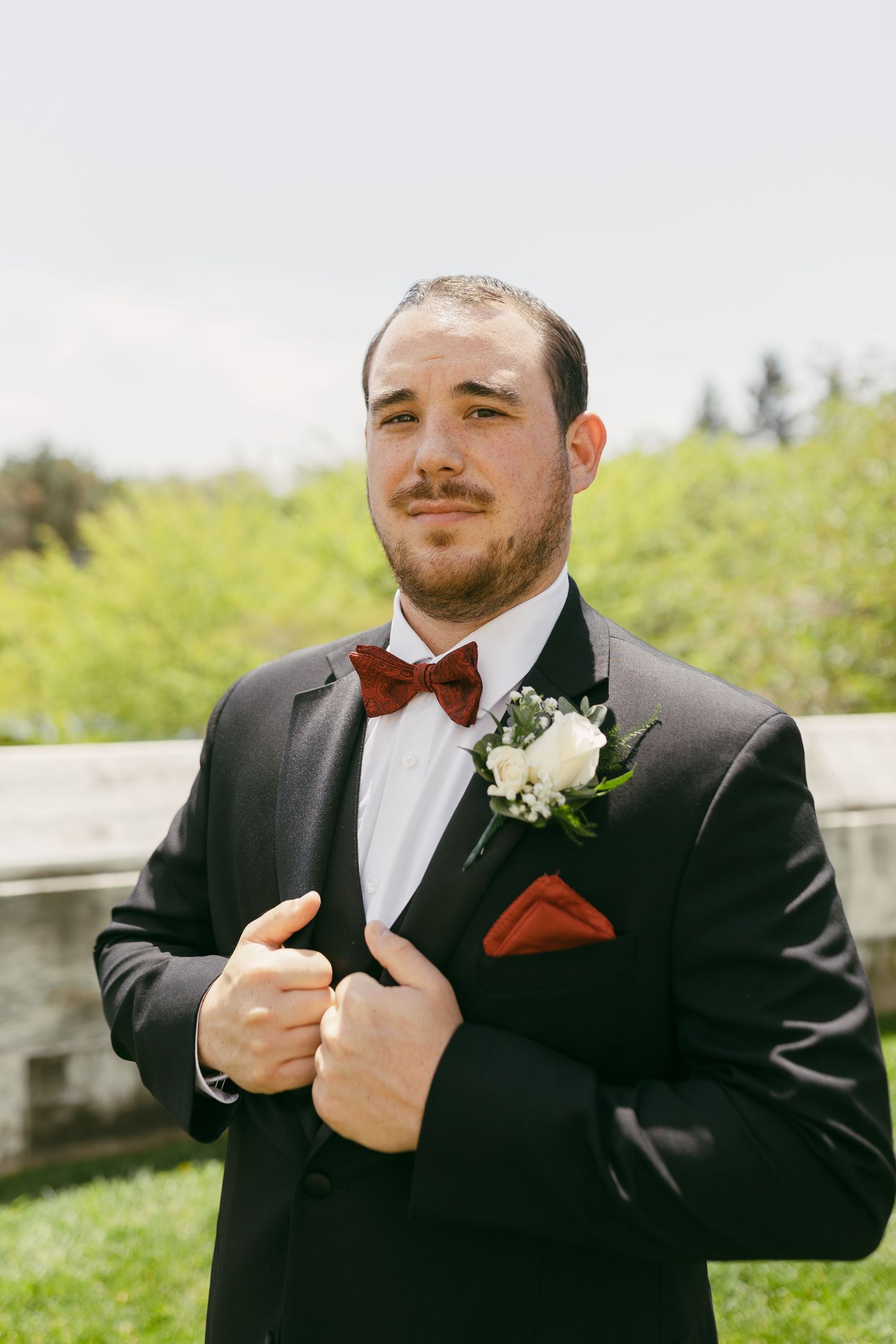Groom in black tuxedo with burgundy bow tie, pocket square, and white boutonniere posing outdoors.