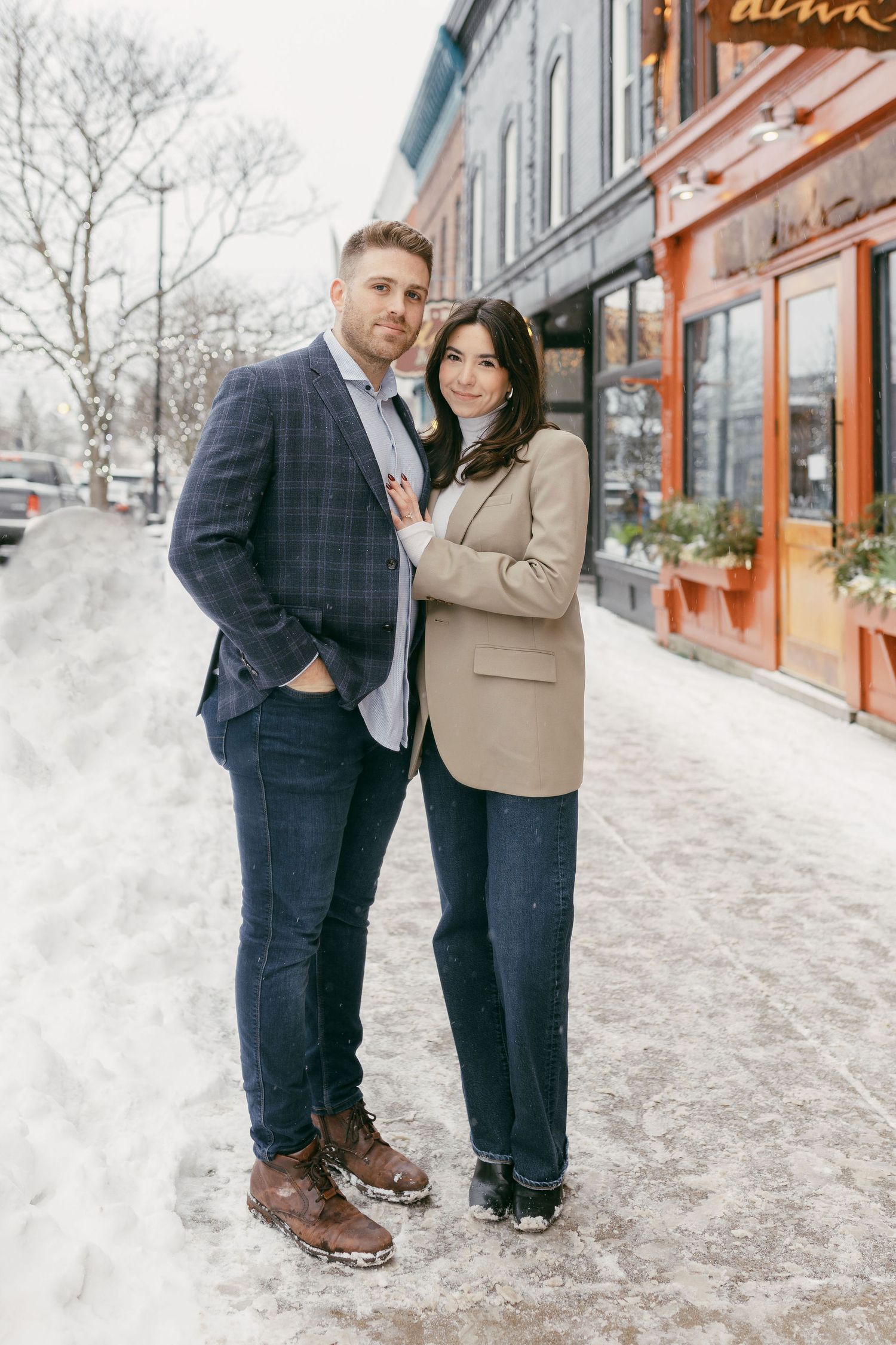 Couple posing together on a snowy sidewalk outside a shop, man in plaid blazer and woman in beige jacket.