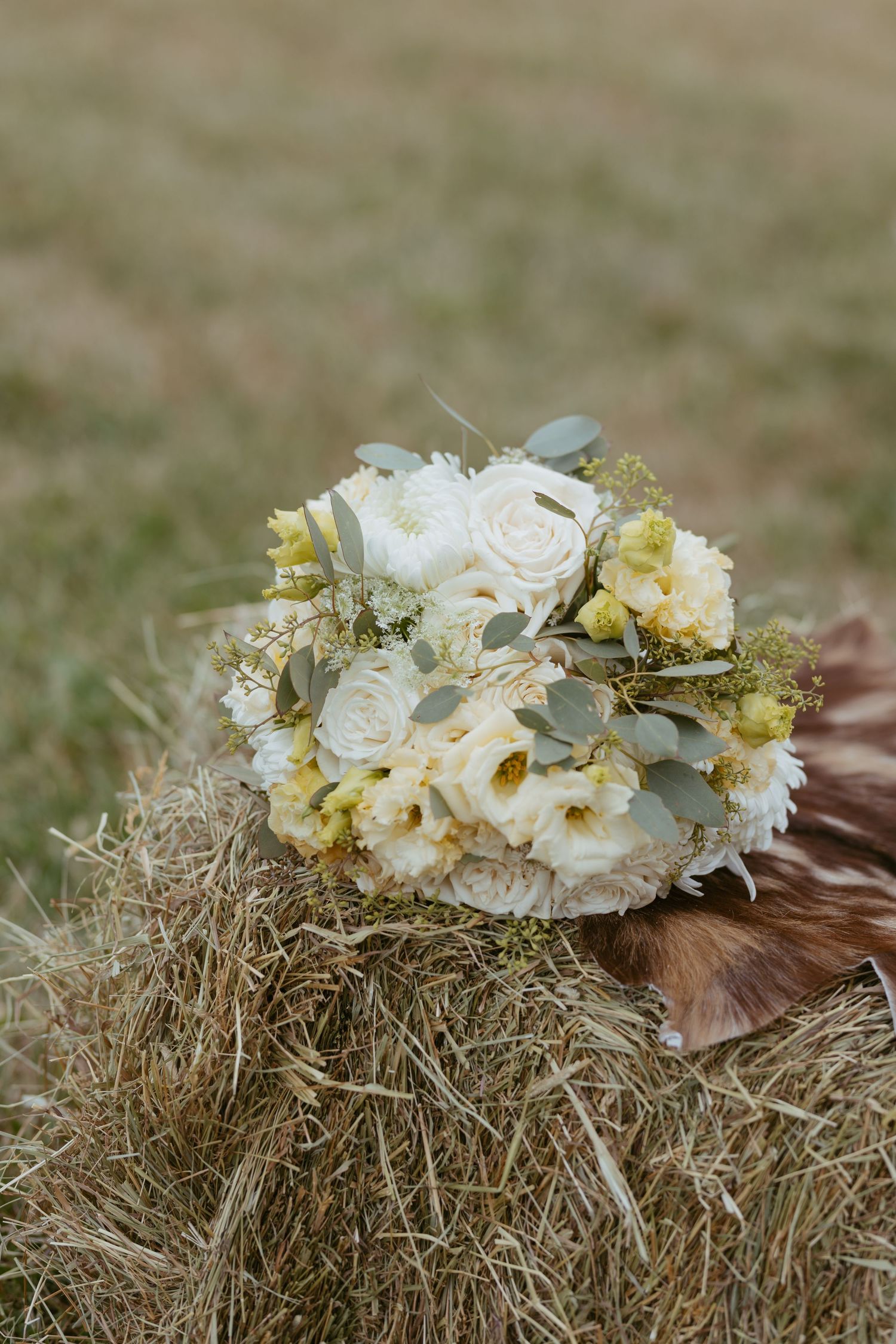 Rustic bridal bouquet with white roses, cream peonies, and greenery resting on a hay bale in a field.