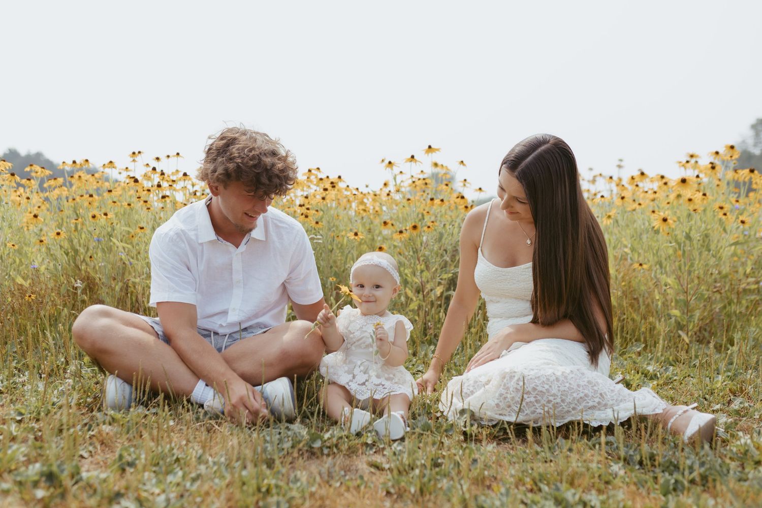 Young family with baby sitting in a sunny wildflower field, parents in white outfits enjoying a peaceful outdoor moment.