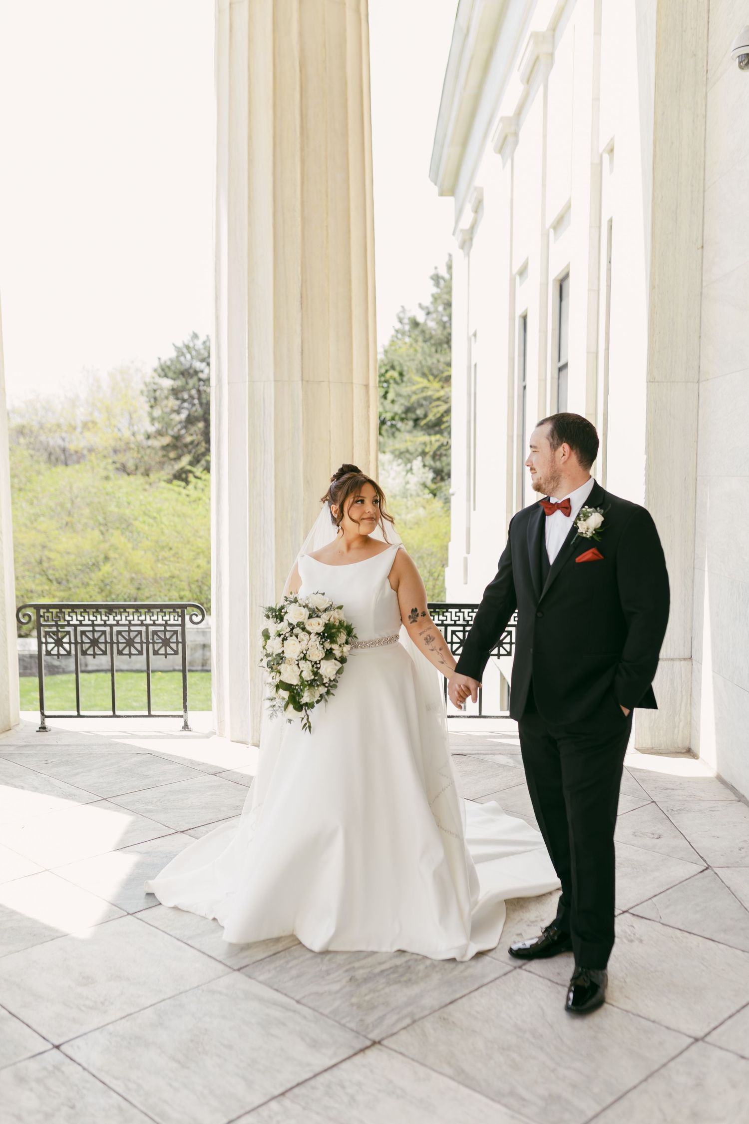 Bride in white gown holding bouquet walks hand-in-hand with groom in black tuxedo on elegant columned venue terrace.