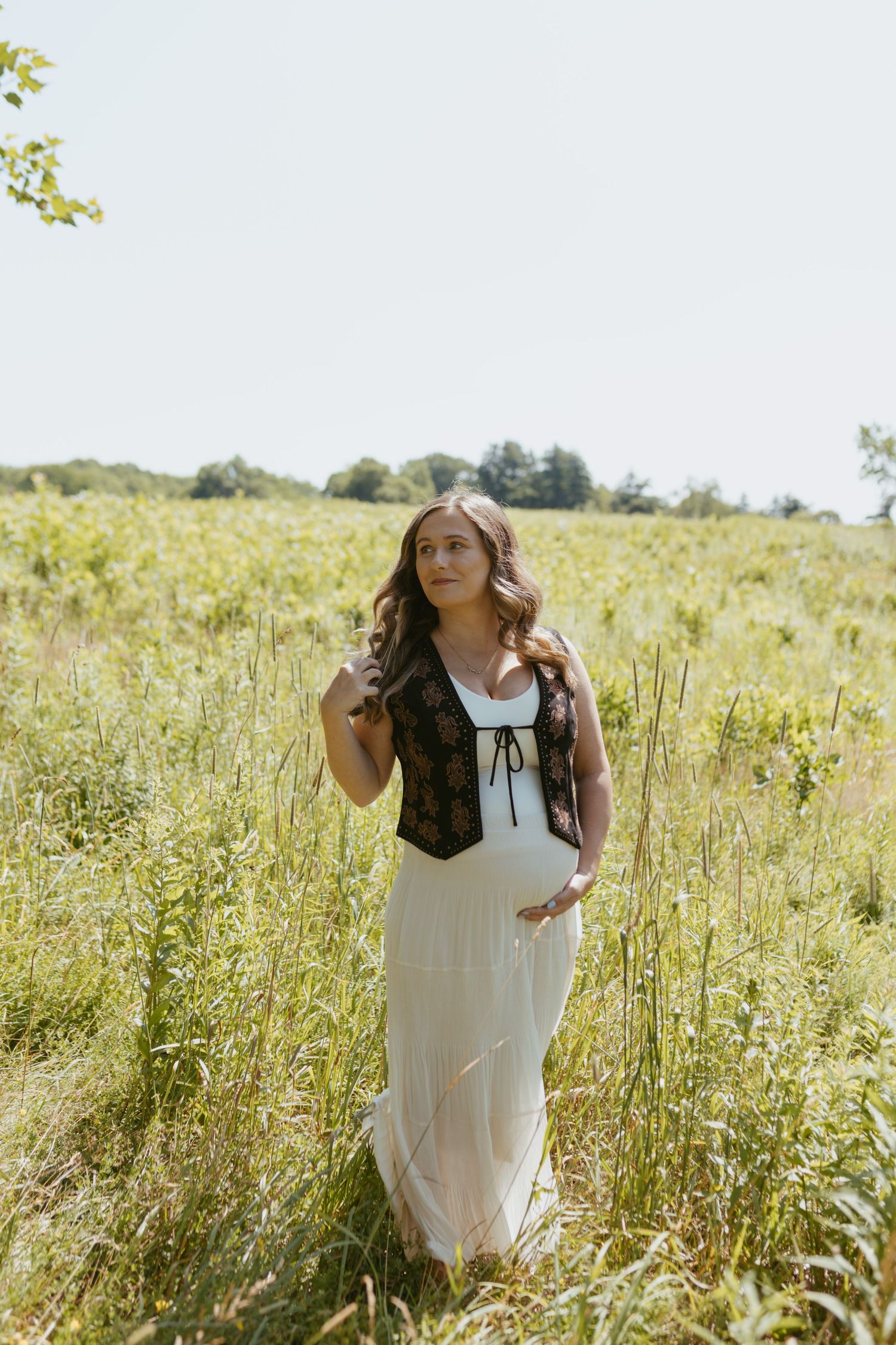 Pregnant woman in white dress and floral corset standing in a sunny green meadow field.