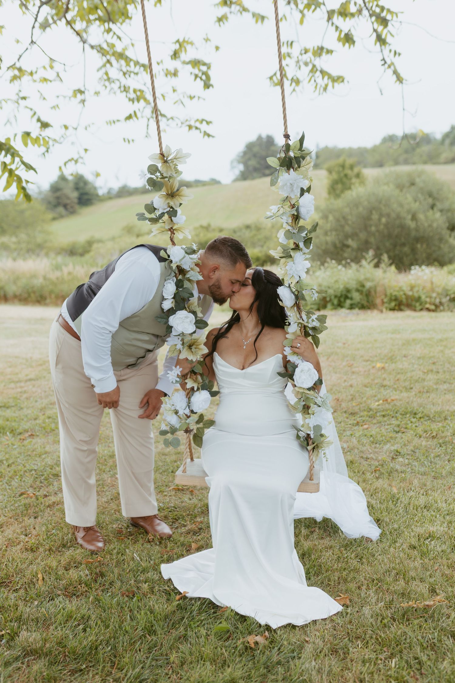 Bride sitting on a floral swing being kissed by groom in tan suit at outdoor wedding.