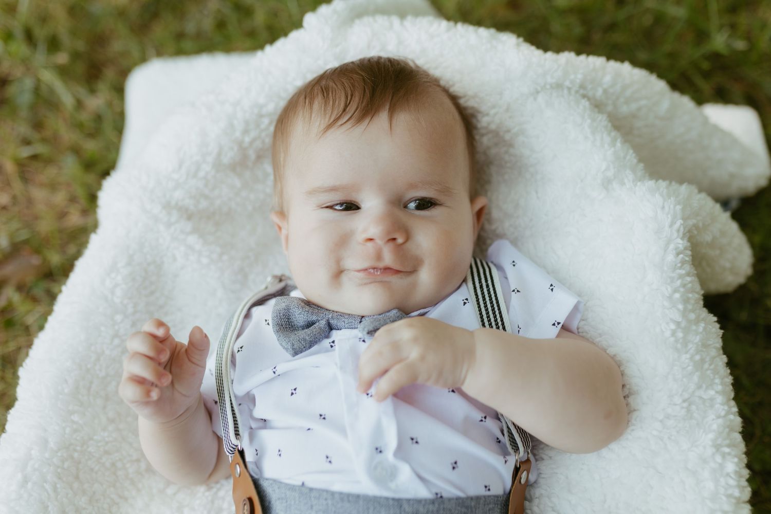 Smiling baby boy in a bow tie onesie lying on a white blanket outdoors on green grass.
