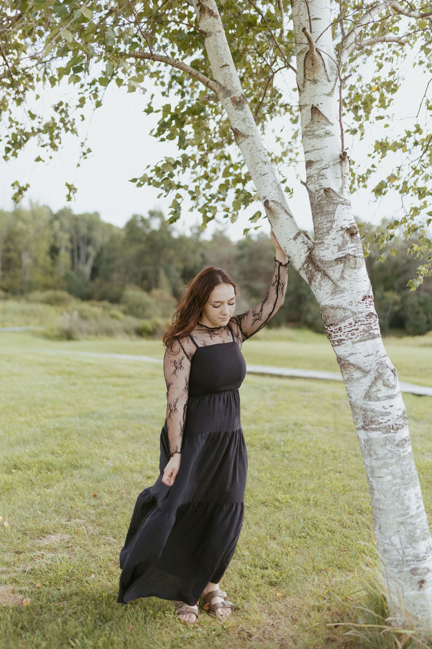 Young woman in black maxi dress with lace sleeves poses gracefully by a birch tree in a lush green park.