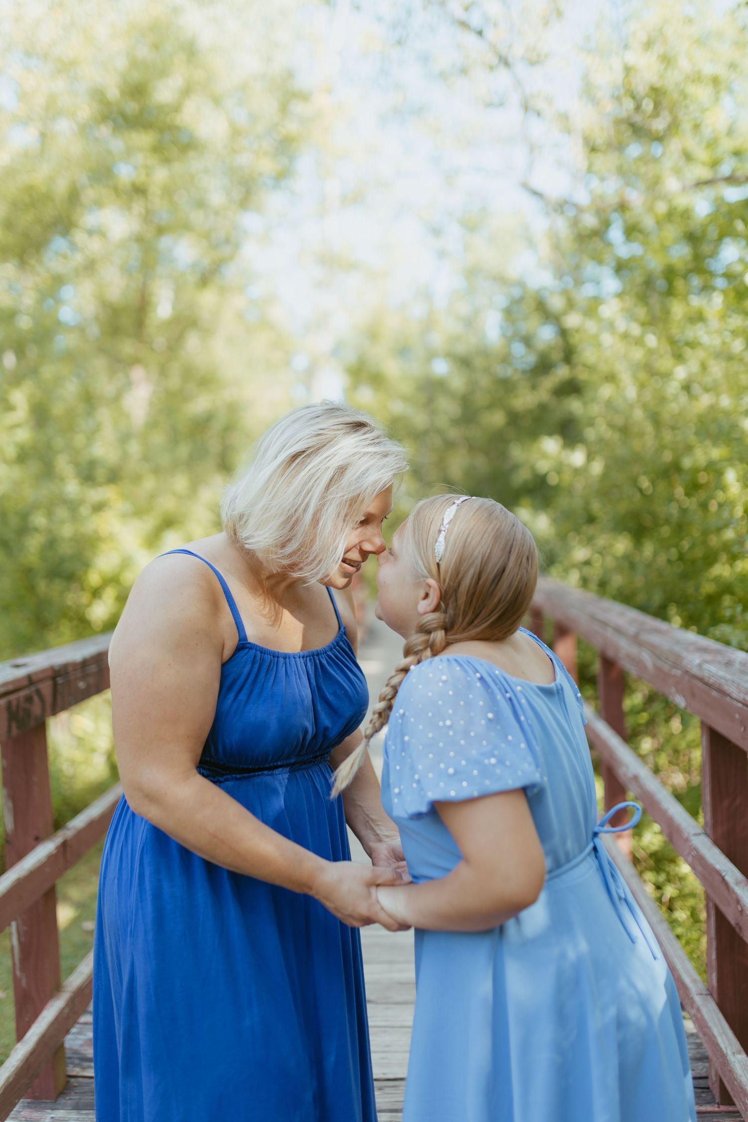 A woman and young girl in matching blue dresses share a tender moment on a wooden bridge surrounded by green trees.