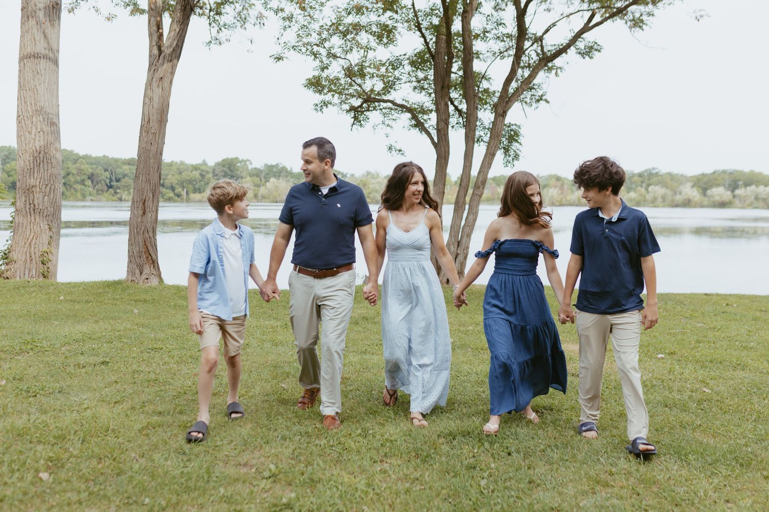 Family of five walking hand-in-hand outdoors near a lake, dressed in coordinated blue and neutral outfits.