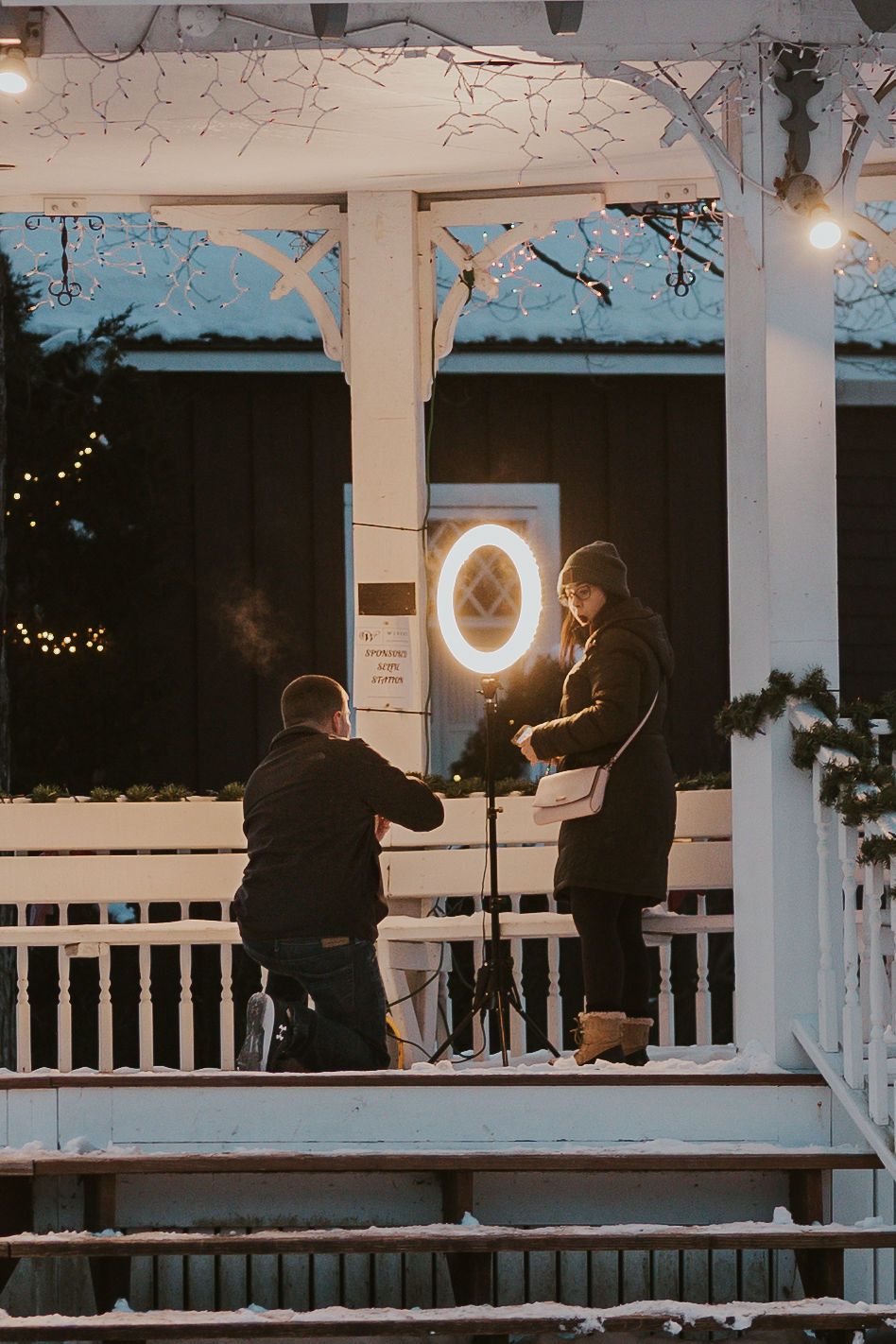Man proposing to woman on snowy decorated porch with ring light, fairy lights, and holiday greenery at dusk.