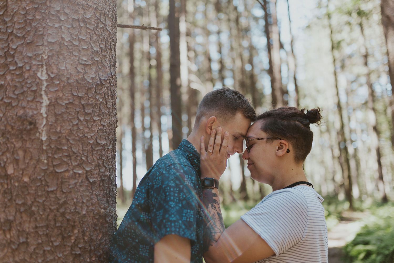 Two men share a romantic moment leaning against a tree in a sunlit forest setting.