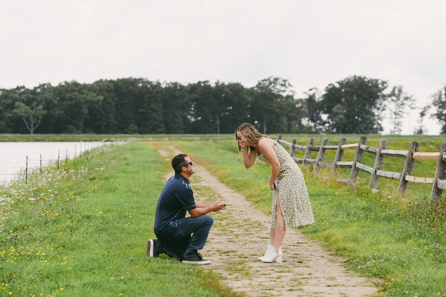 Man kneeling on a garden path proposing to a woman in a floral dress beside a wooden fence in a green field.