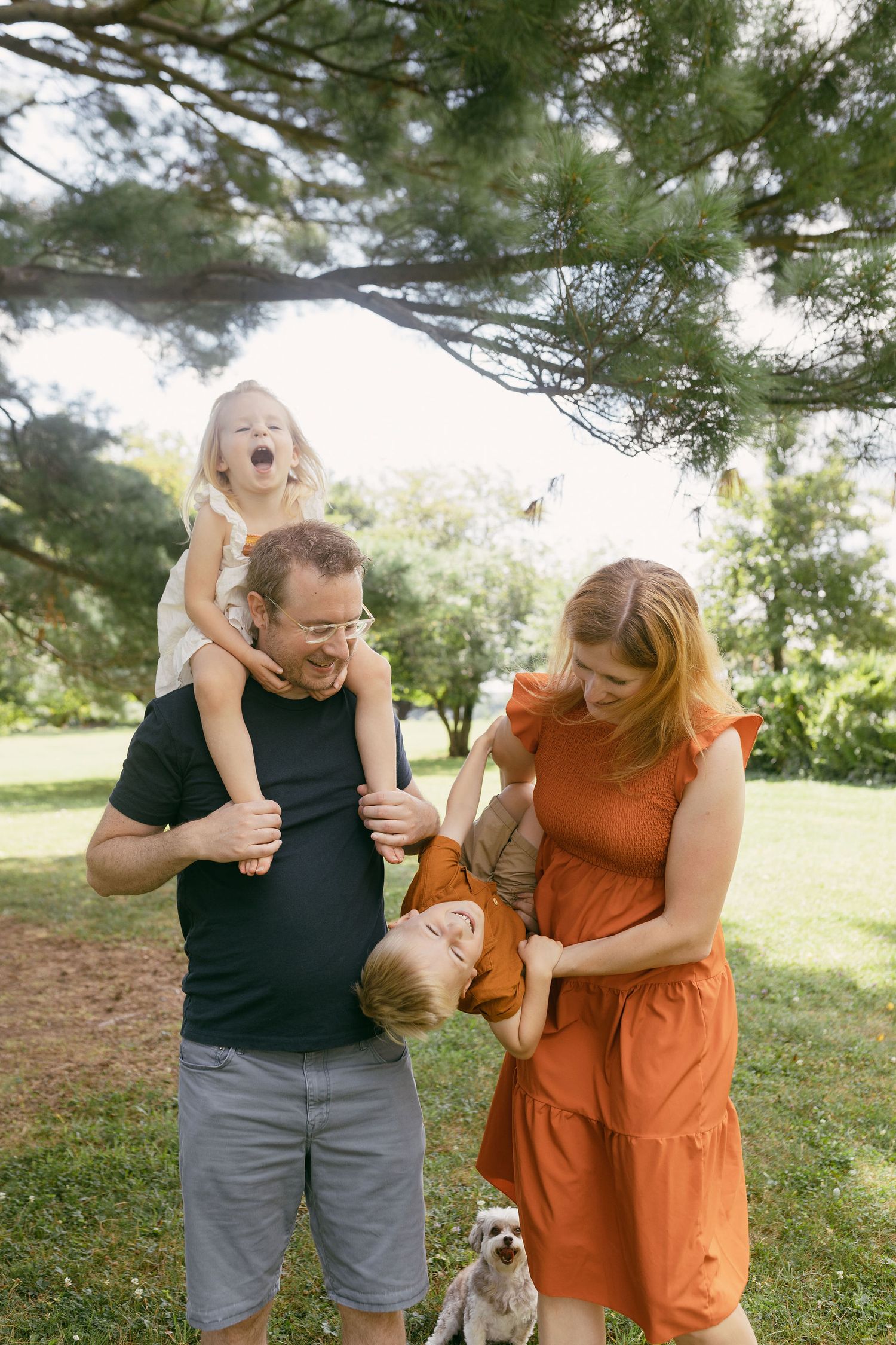 Family of four playing outdoors under pine tree, parents laughing with two young children, small dog nearby on green lawn.