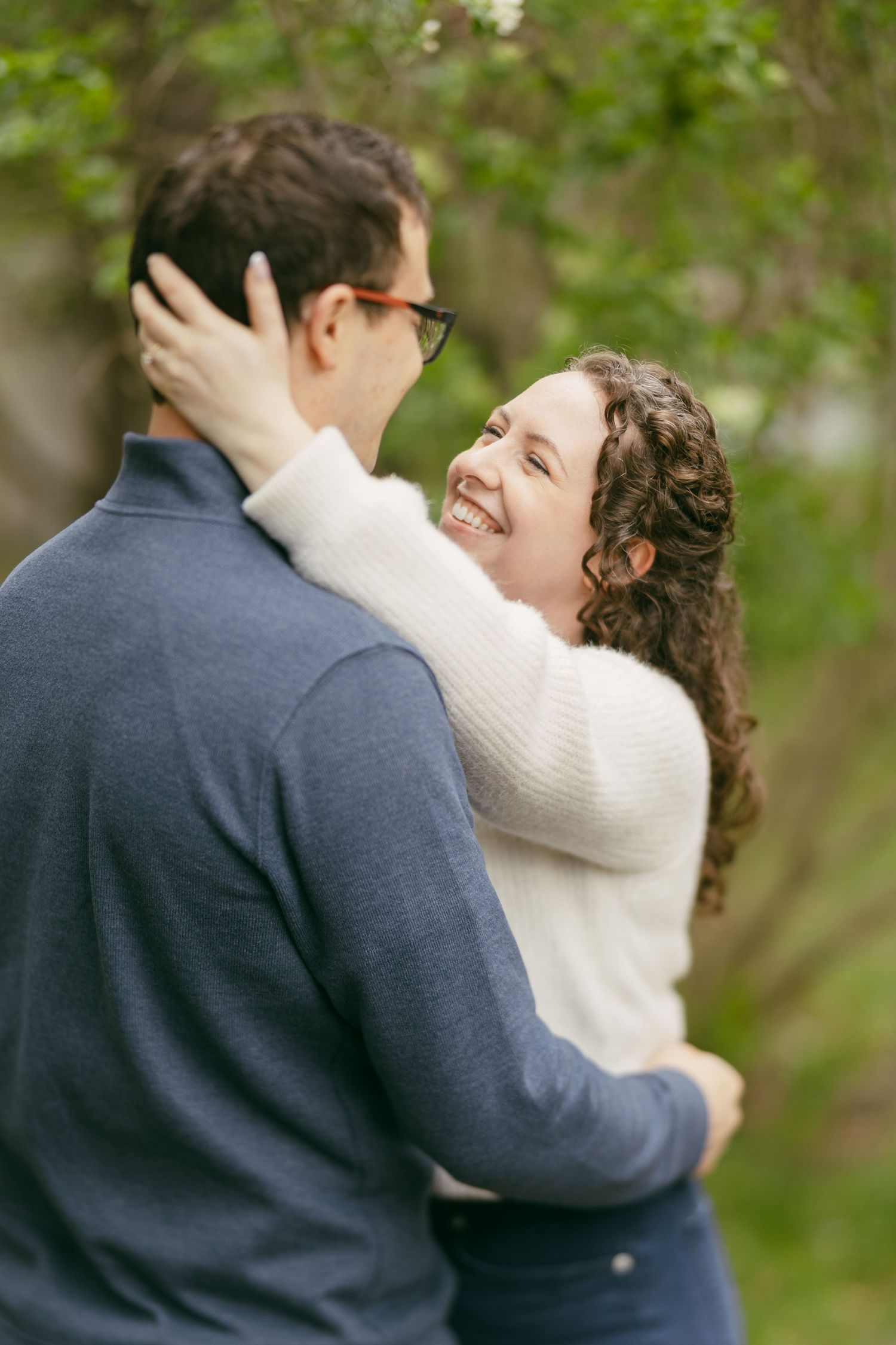 A smiling woman with curly hair embraces a man wearing glasses outdoors among green trees.