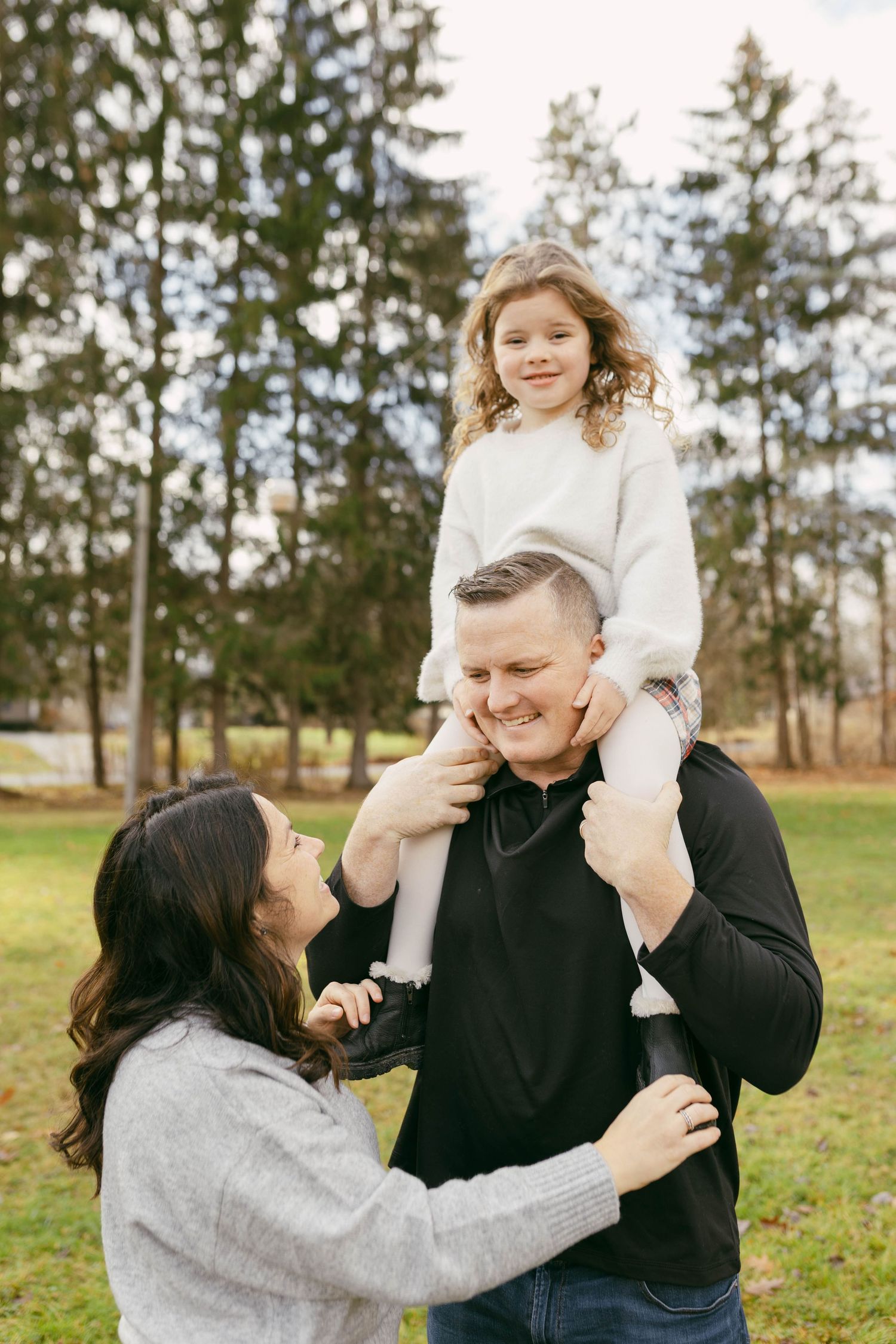 A happy family with a young girl sitting on her father's shoulders outdoors in a park surrounded by tall pine trees.