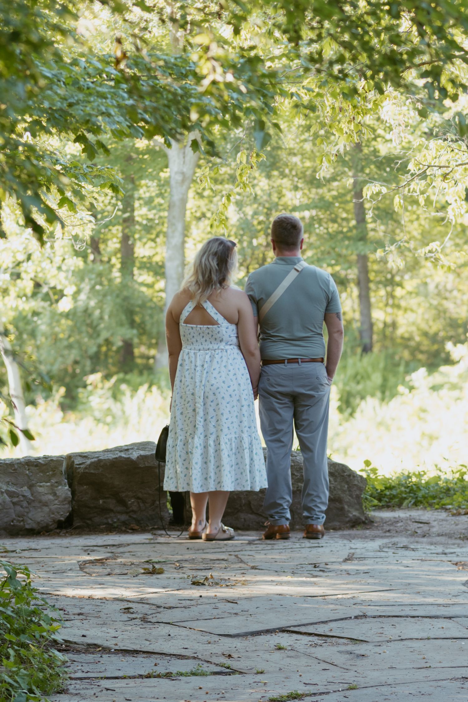 Couple holding hands from behind, standing on stone path surrounded by lush green trees in a park.