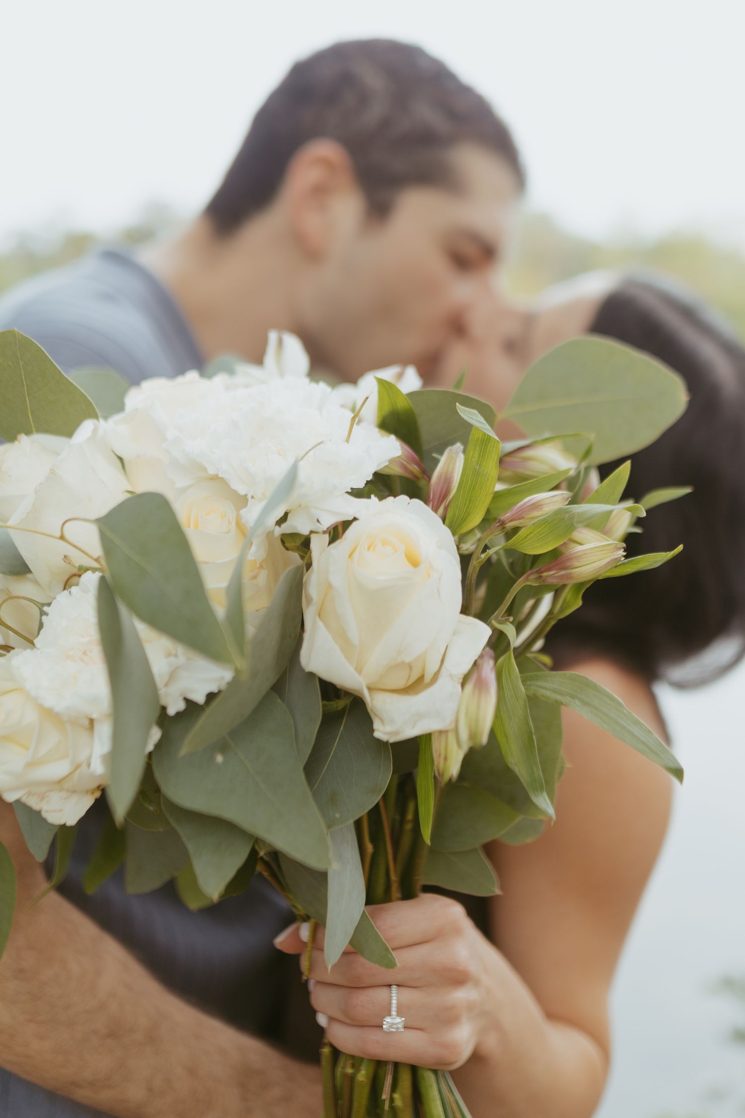 Couple kissing behind a bridal bouquet of white roses and eucalyptus, with an engagement ring visible on her hand.