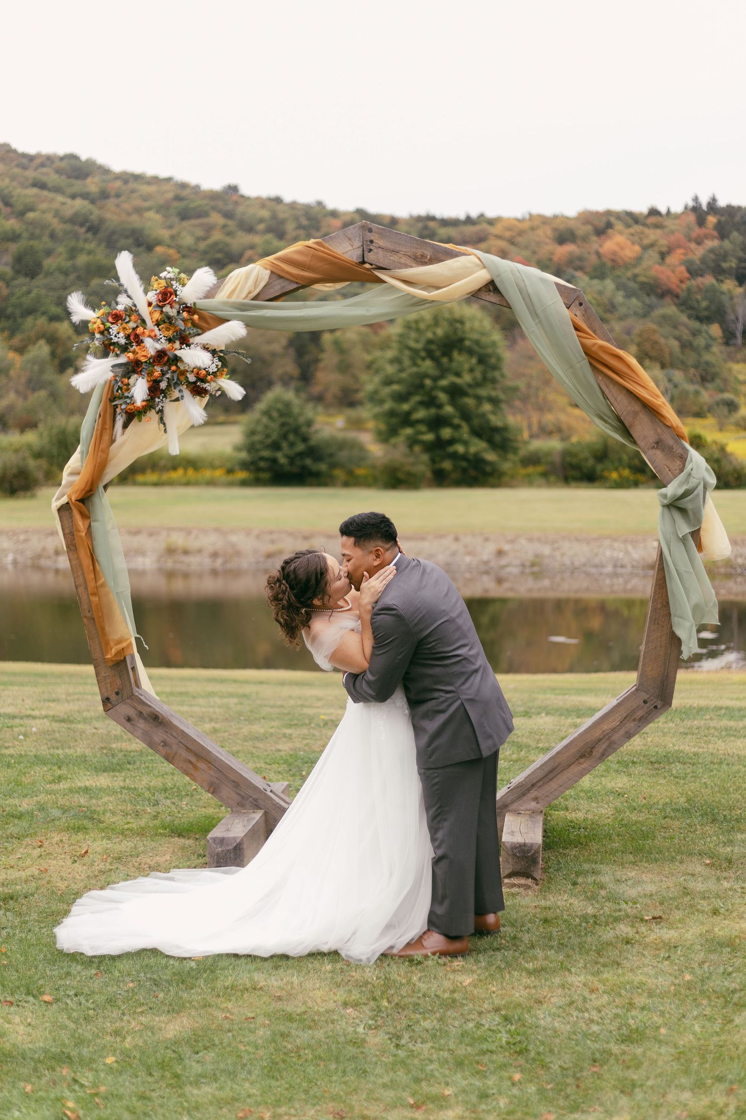 Newlyweds share a kiss under a wooden hexagonal arch by a lake during autumn.
