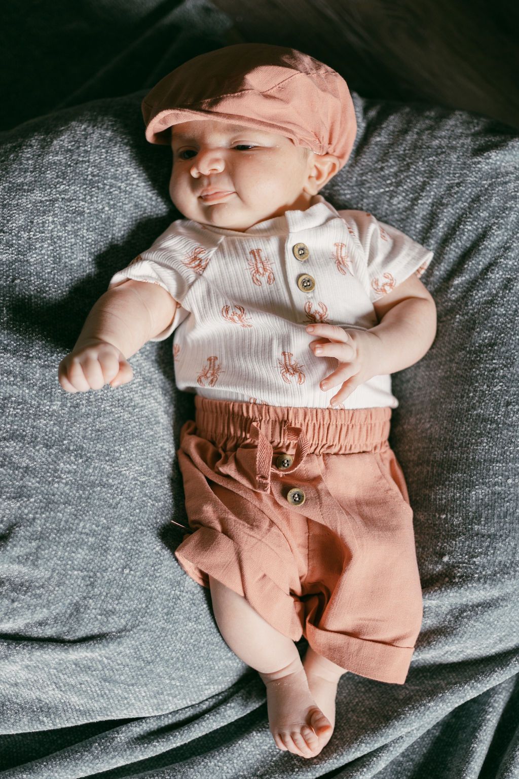 Newborn wearing white top and peach pants lying on gray fabric.