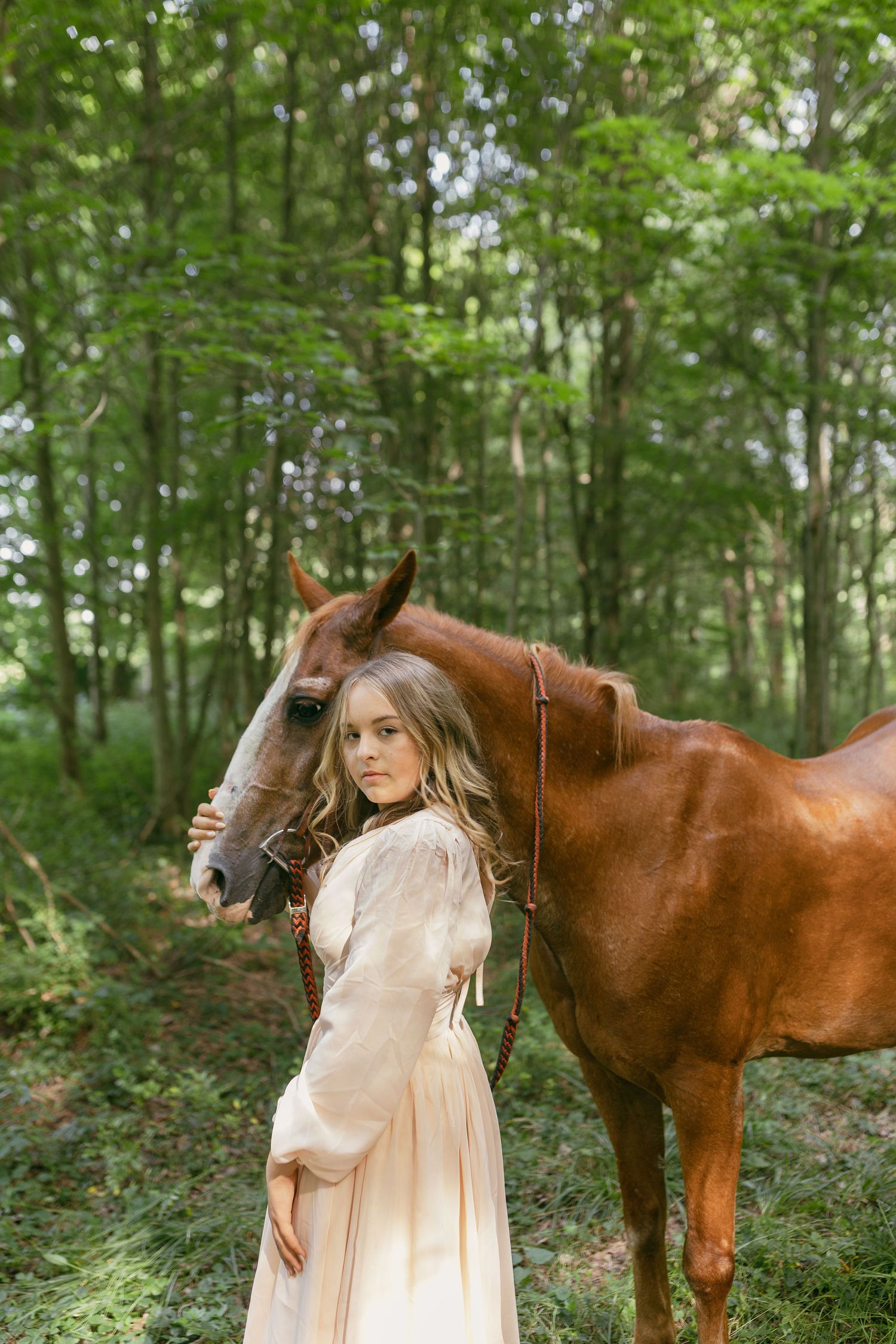 A brown horse and person in a light-colored dress pose together in a lush woodland setting.