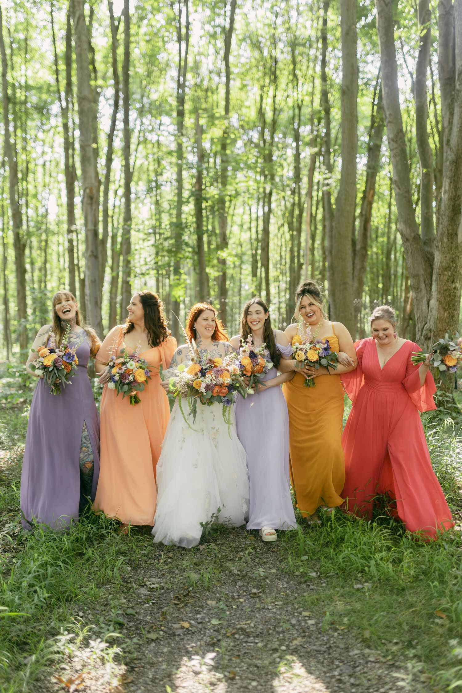 Bridesmaids in colorful coral, purple, and mustard dresses pose with bouquets in a sunlit forest setting.