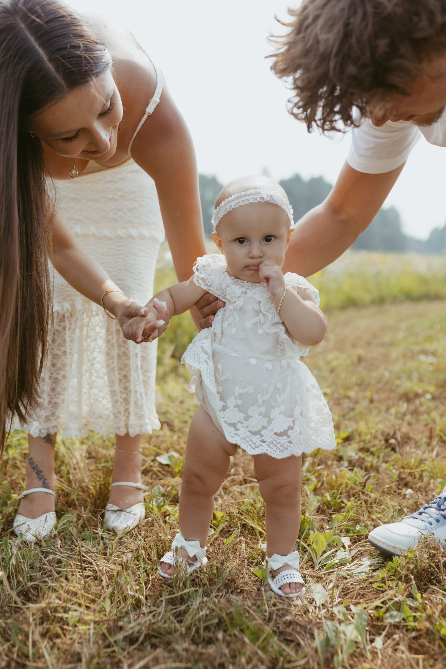 Parents helping baby take first steps outdoors in matching white outfits on a sunny day in a green field.