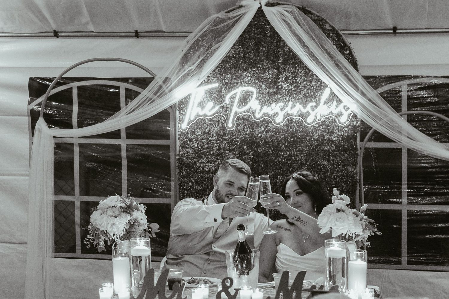 Bride and groom toast at wedding reception sweetheart table with neon sign in black and white photo.