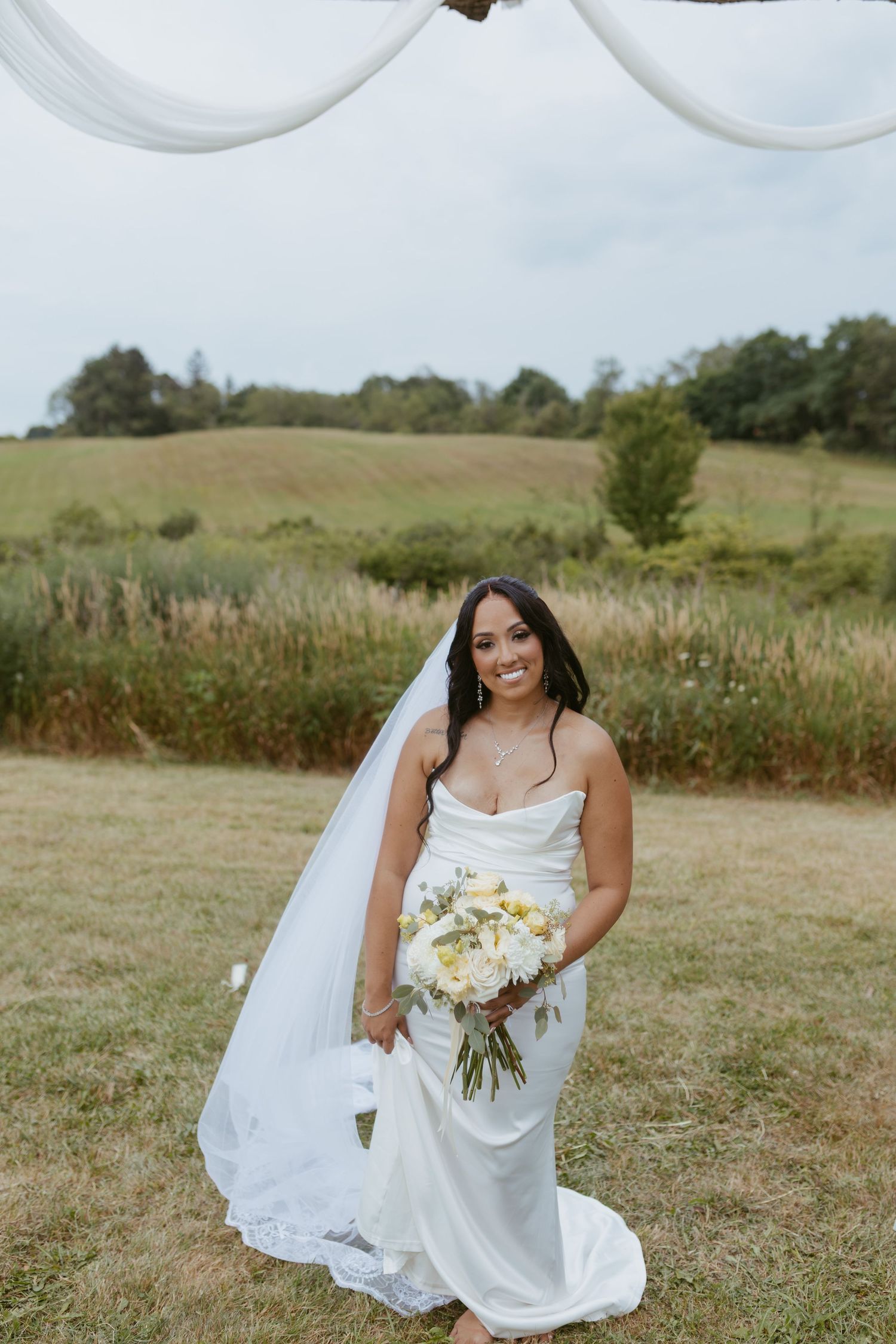 Smiling bride in white fitted gown holds bouquet at outdoor wedding with rolling green fields behind her.