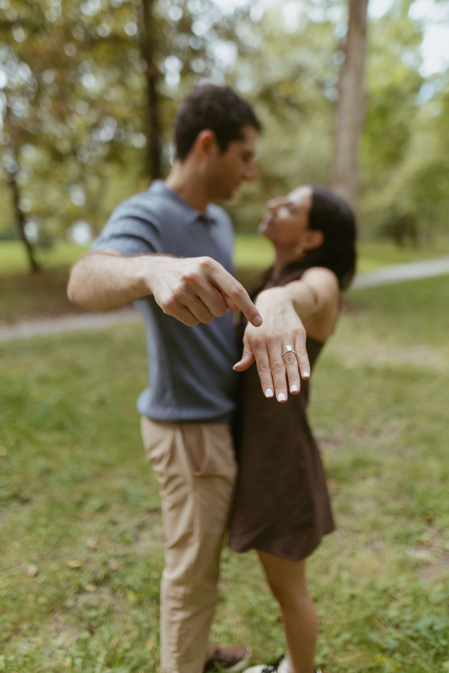 Couple in a park showing off engagement ring, smiling and leaning in for a kiss outdoors.