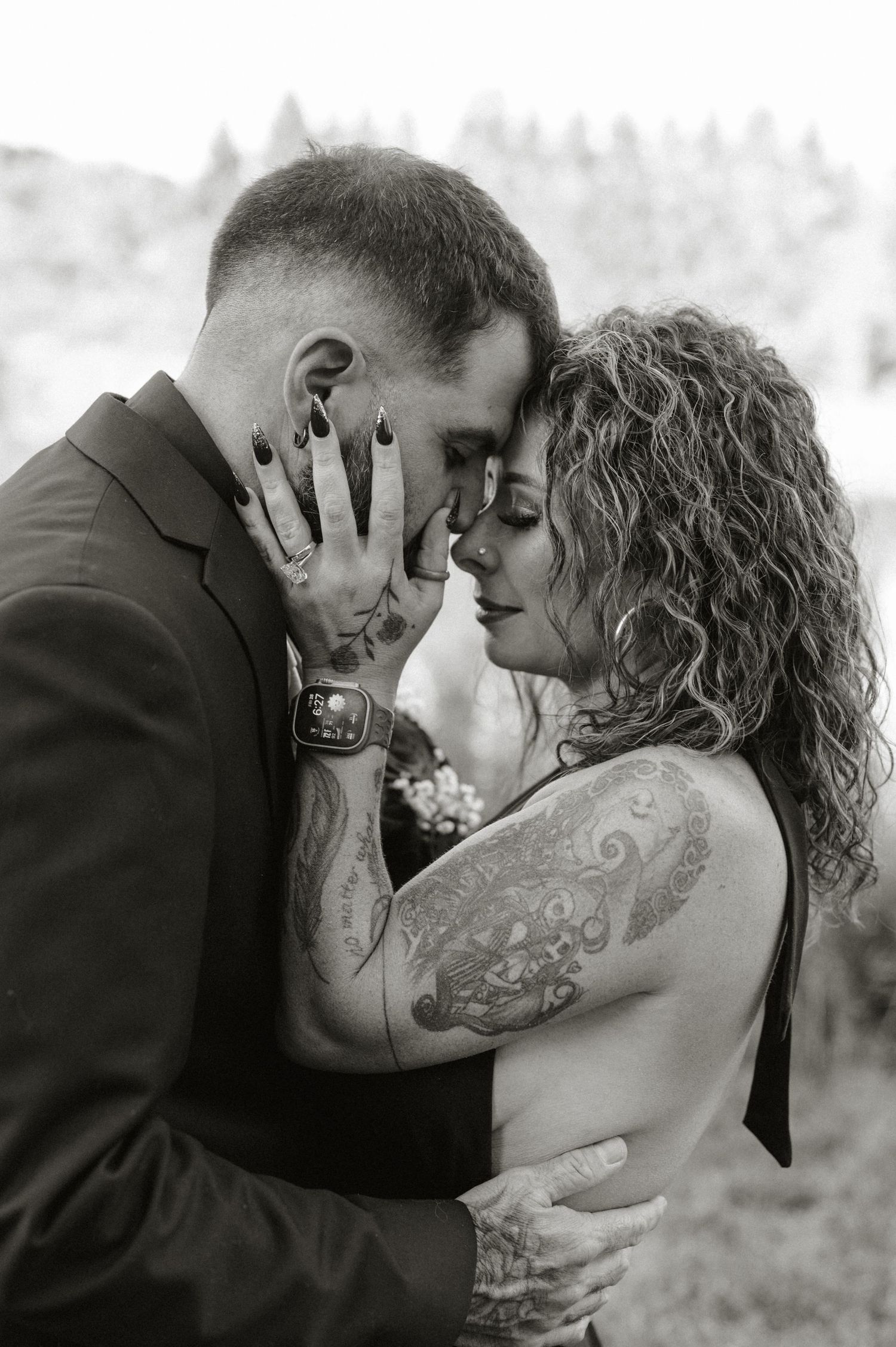 Black and white photo of tattooed couple in wedding attire sharing an intimate forehead touch embrace outdoors.