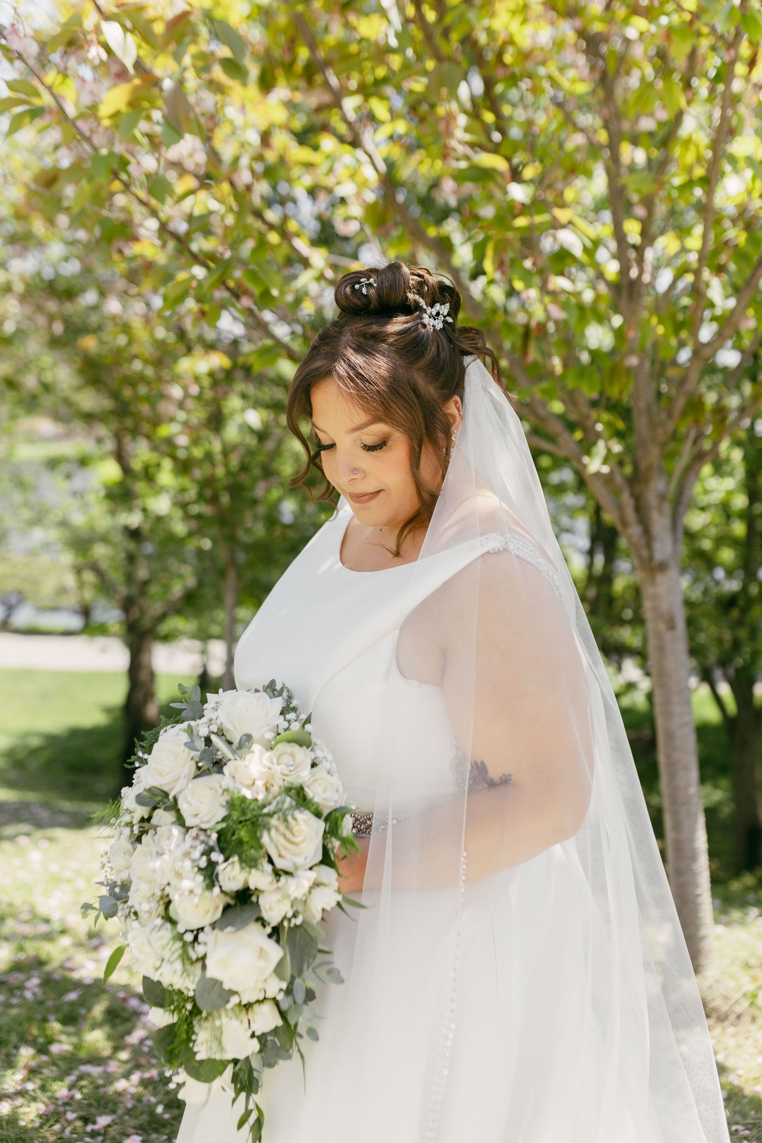 Bride in white gown and veil holding cascading white rose bouquet, standing among green trees.