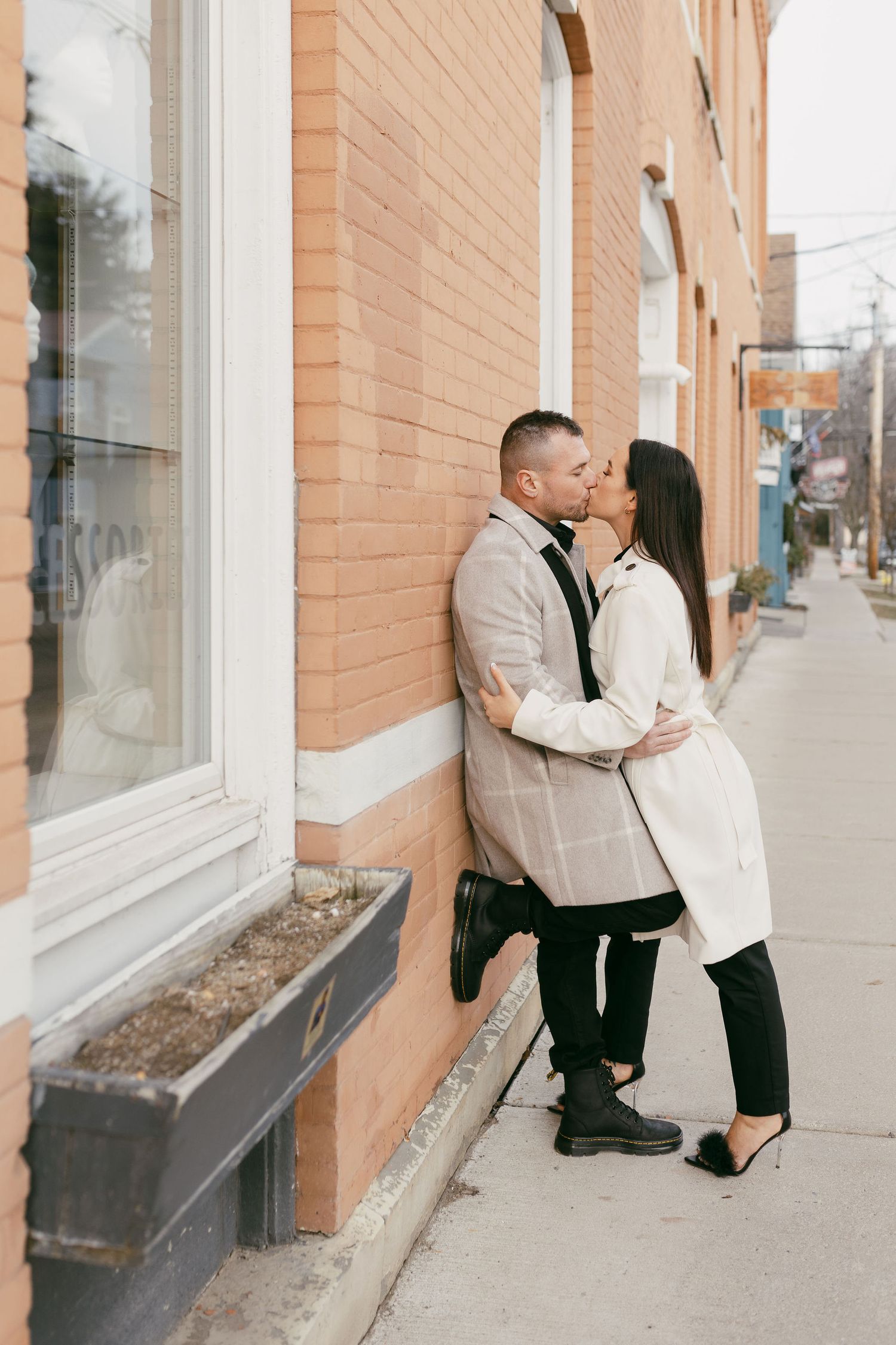 Couple sharing a romantic kiss against a brick building wall on a city sidewalk, dressed in stylish coats.