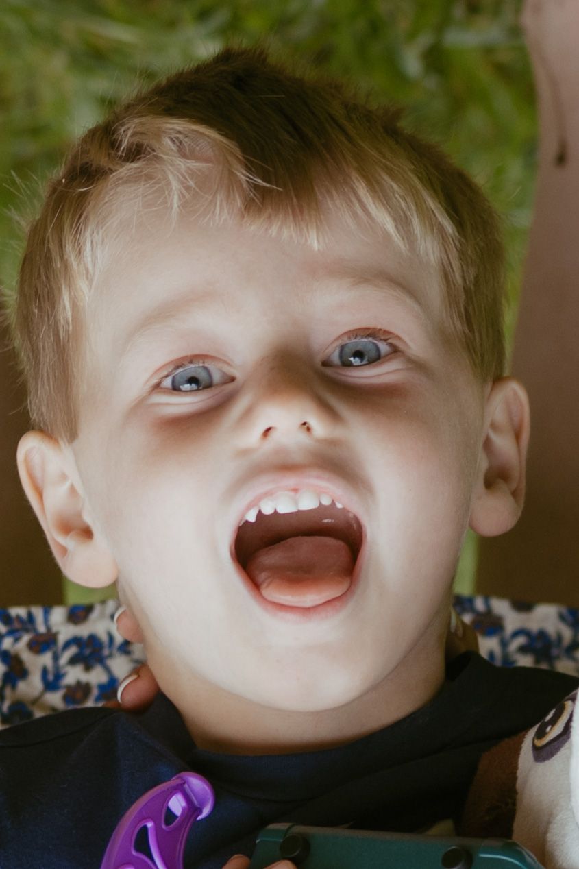 A young toddler boy with blue eyes and blonde hair laughs with mouth wide open outdoors.