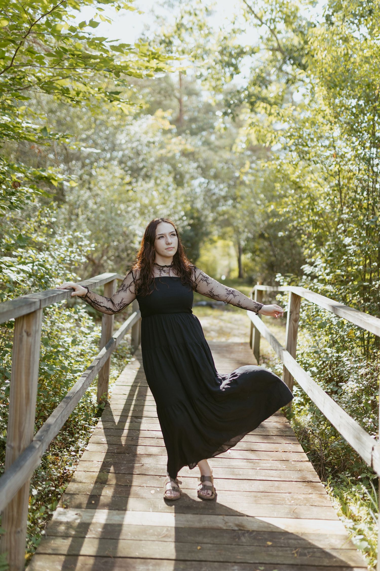 Young woman in black maxi dress posing on wooden bridge surrounded by lush green trees in sunlit forest.
