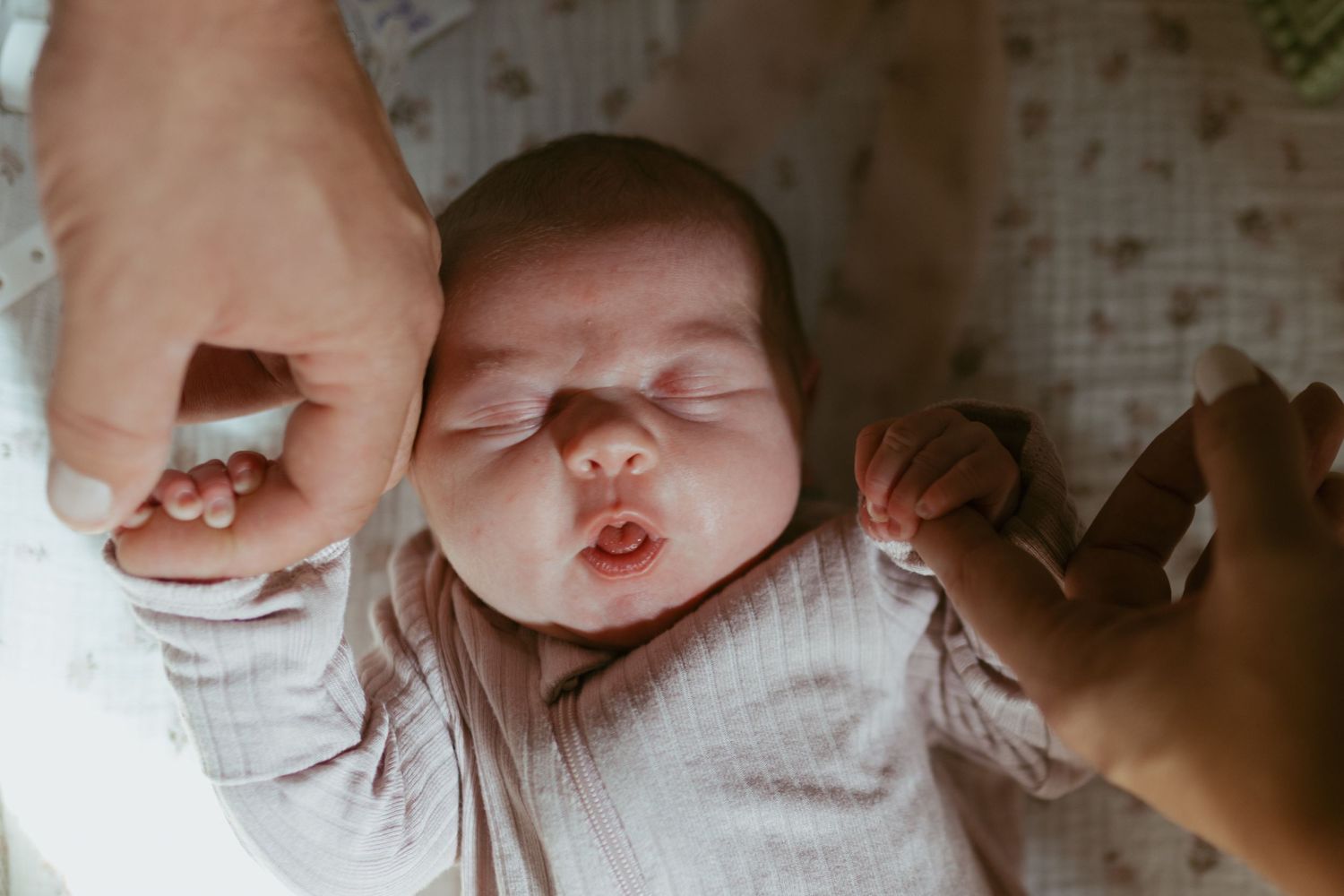 Newborn baby lying down holding parents fingers with eyes closed in a peaceful nursery setting.