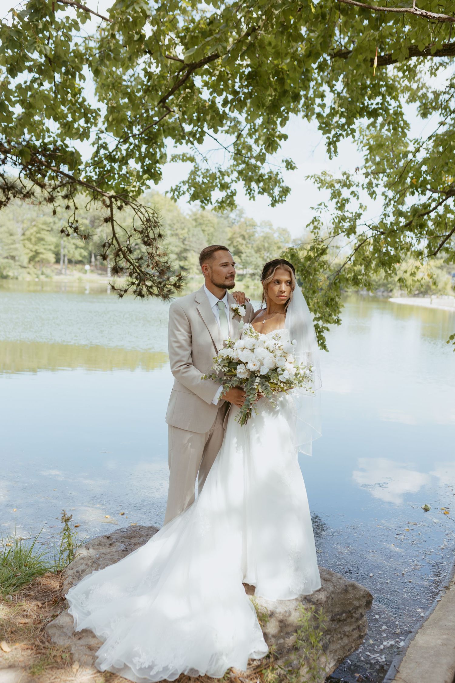 Bride and groom pose by a serene lake under lush green trees, bride holding a white floral bouquet in elegant wedding attire.